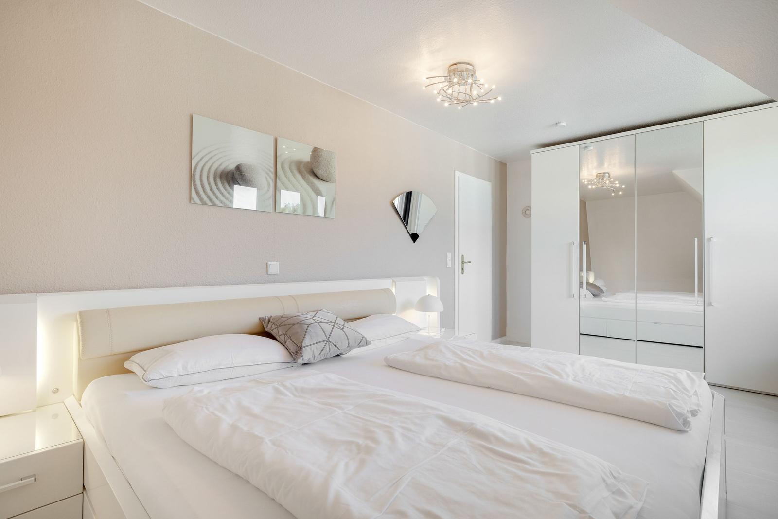 Schlafzimmer mit weißem Bett, Kleiderschrank und Wandbildern.