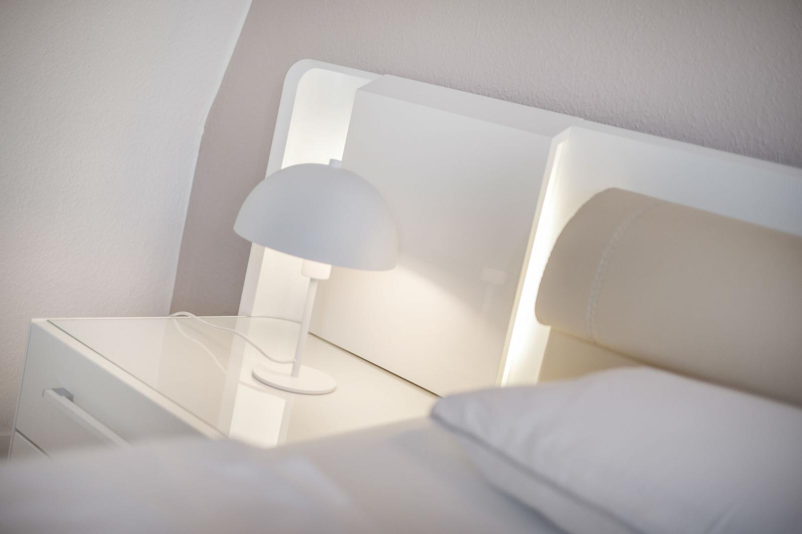 Schlafzimmer mit weißem Bett, Nachttisch und Lampe.