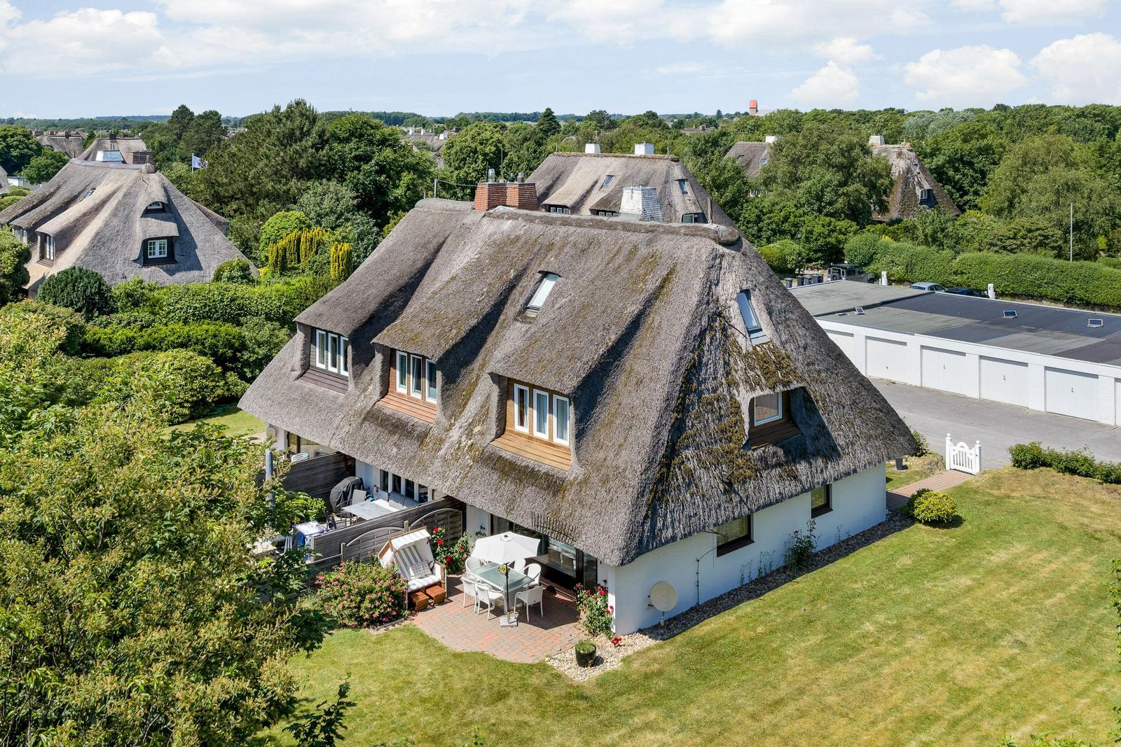 Haus mit Strohdach, Terrasse und Garten. Umgeben von Bäumen und Grünflächen.