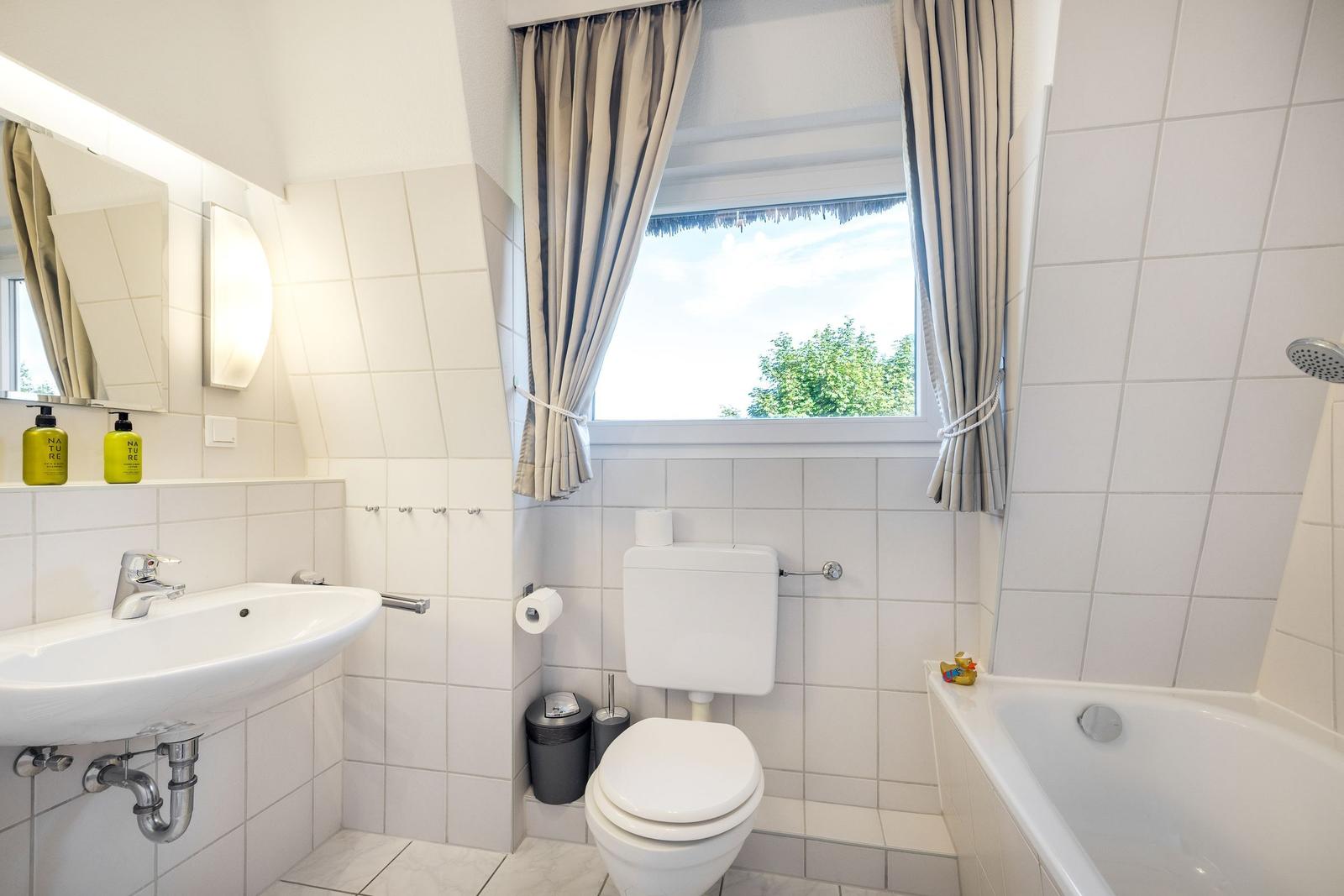 Badezimmer mit Badewanne, Toilette, Waschbecken und Fenster mit Blick auf Grün.