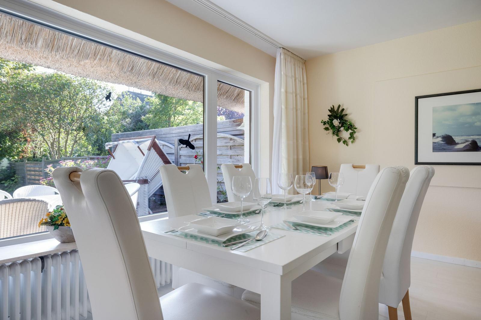 Esszimmer mit weißem Tisch und Stühlen, großes Fenster zum Garten, Wandbild und Blumendekoration.