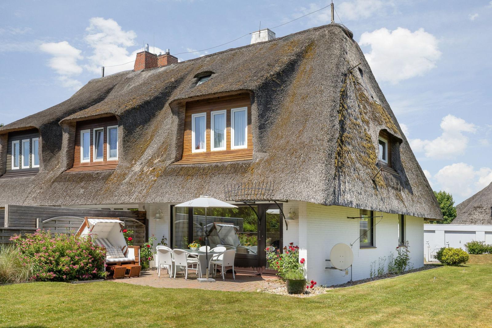 Haus mit Strohdach, Terrasse mit Tisch und Stühlen, Garten mit Blumen und Liegestuhl.