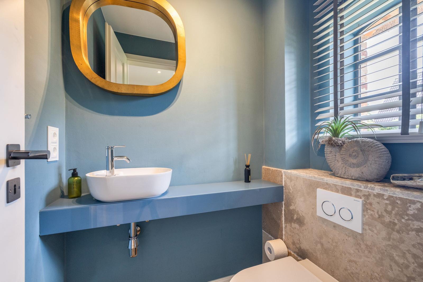 Badezimmer mit Waschbecken, Spiegel und Fenster mit Jalousie.