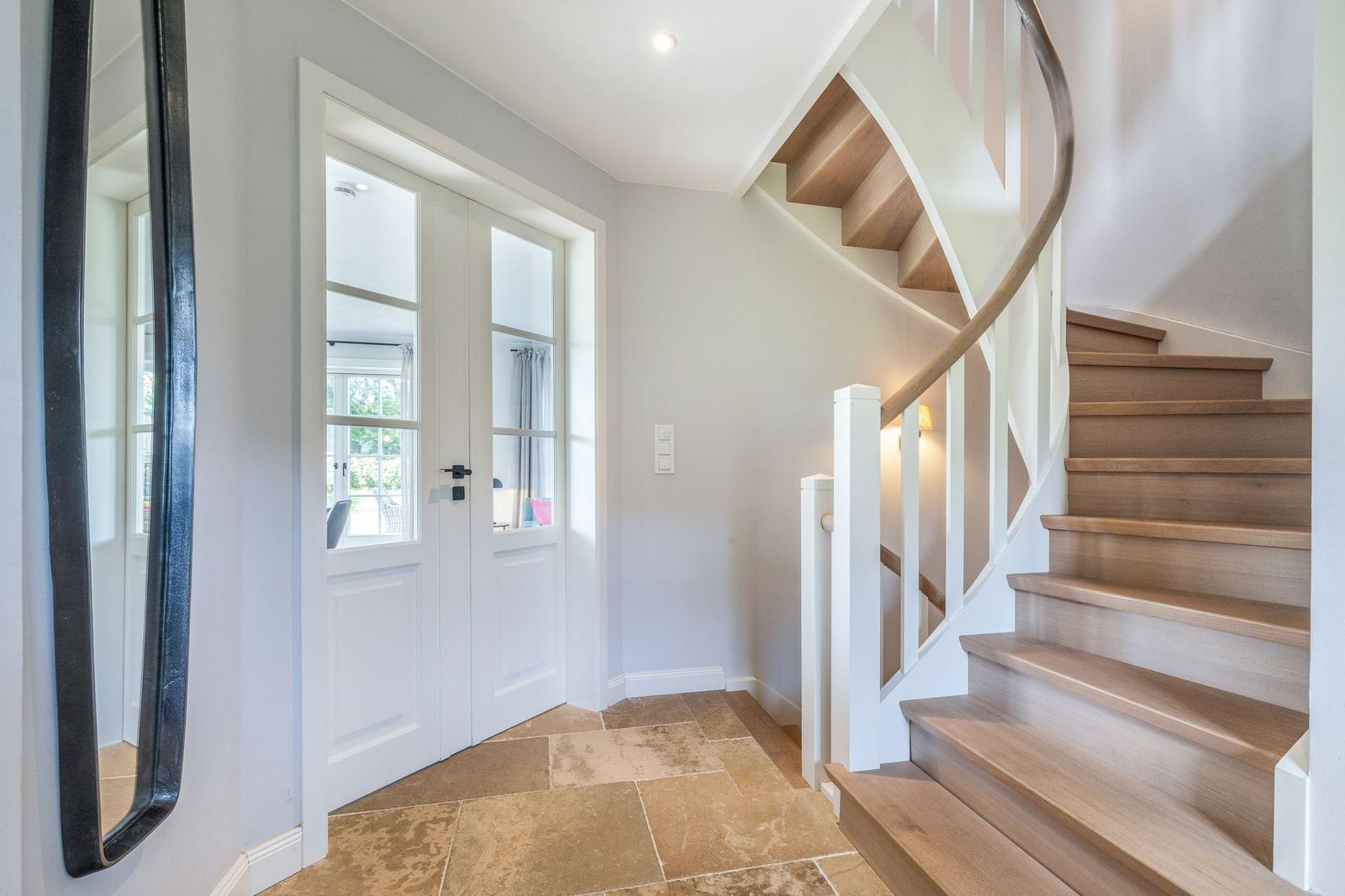 Hausflur mit Treppe und Tür. Holzstufen, weißer Geländer, Steinboden.
