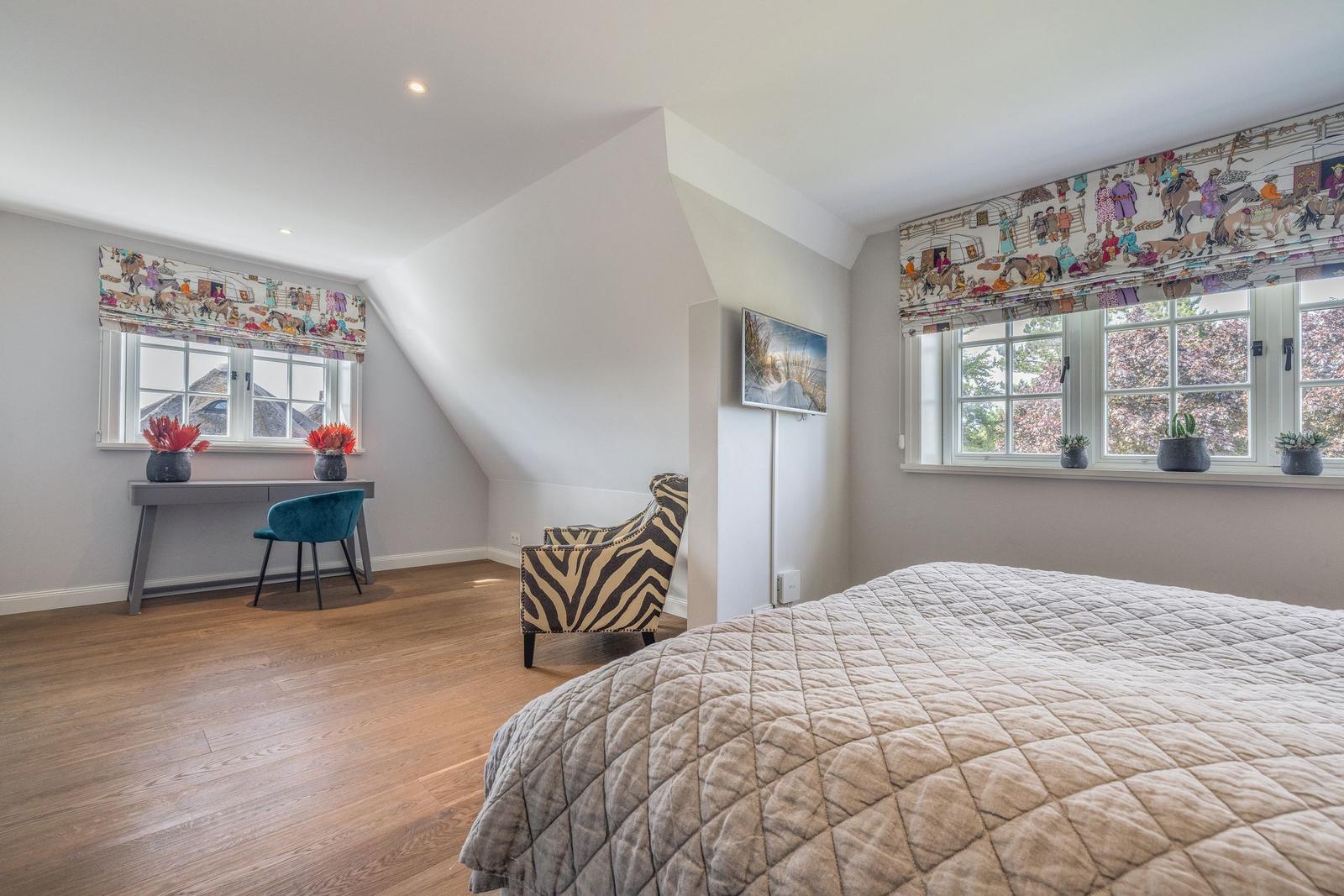 Schlafzimmer mit Dachschrägen, zwei Fenstern, Bett, Schreibtisch und Sessel.