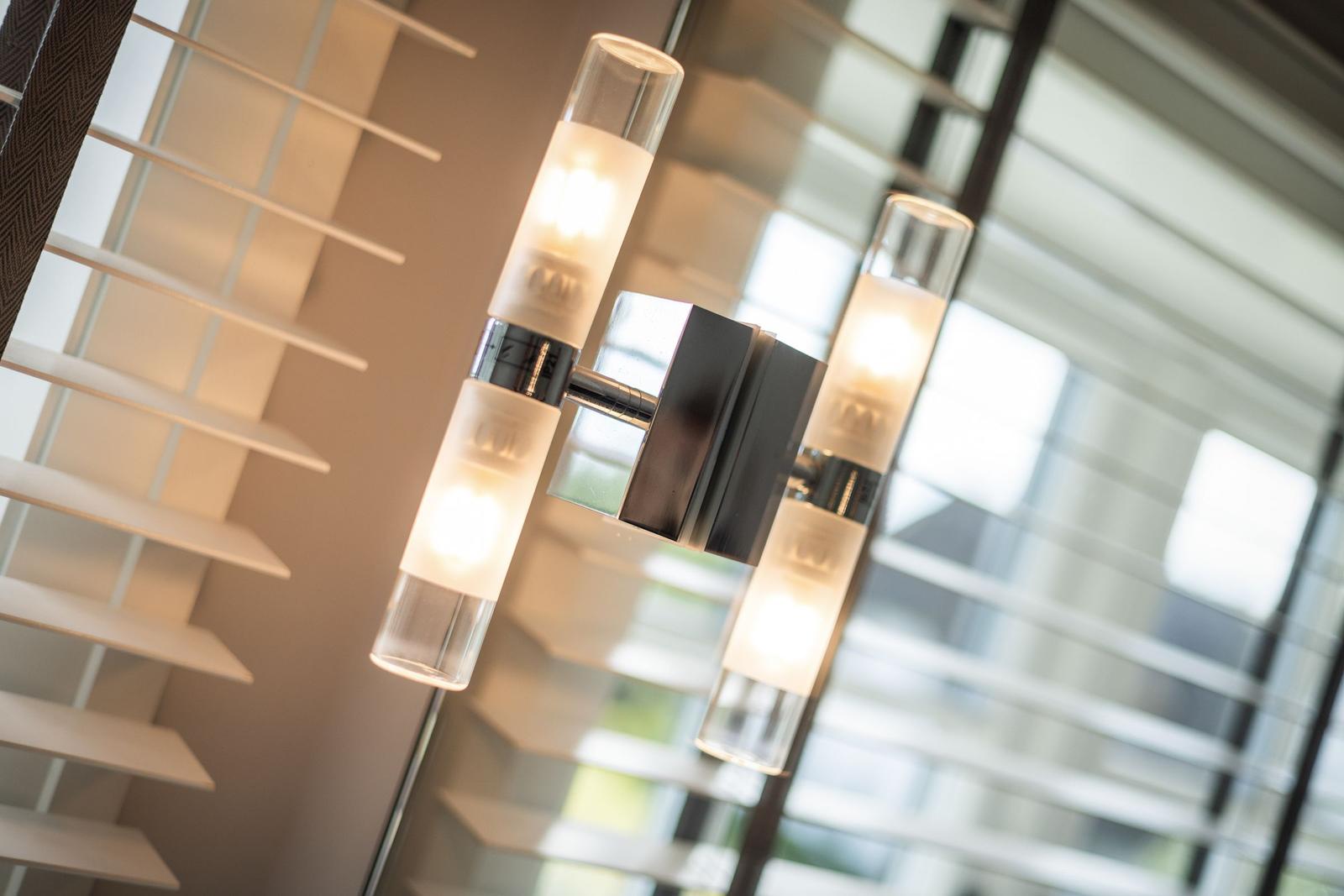 Moderne Wandlampe mit vier Glasröhren und schwarzem Metallgestell vor Jalousien.