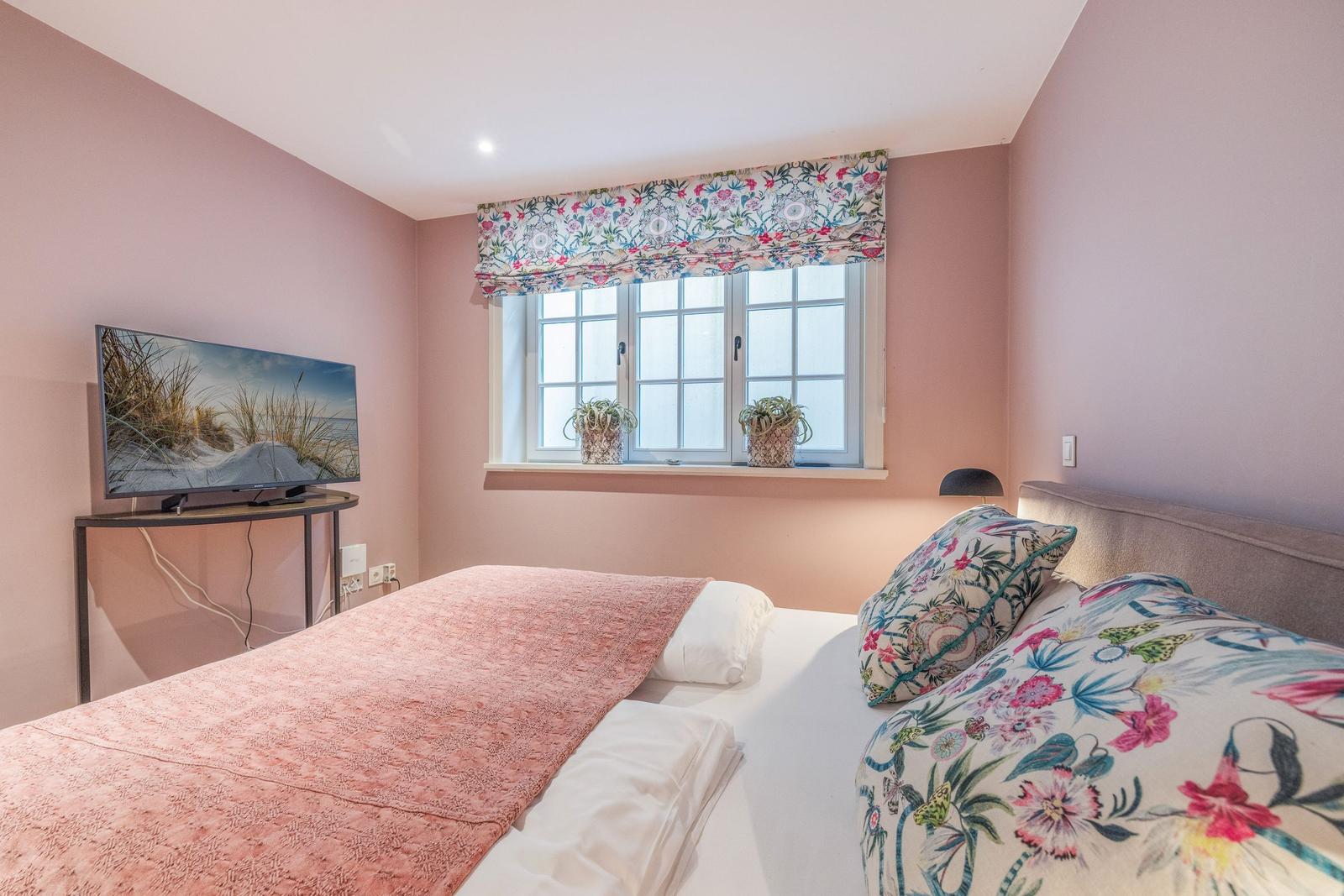 Schlafzimmer mit Bett, Fenster, TV und Blumenstoffen an Wand und Bett.