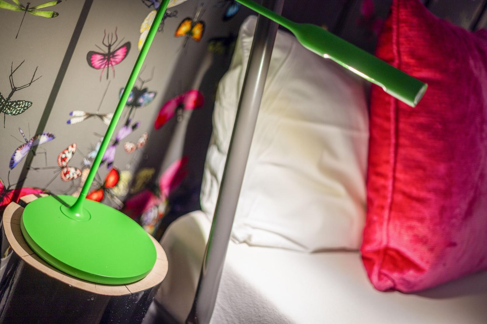 Grüne Lampe auf Nachttisch neben Bett mit Kissen und Wand mit Schmetterlingsmotiven.