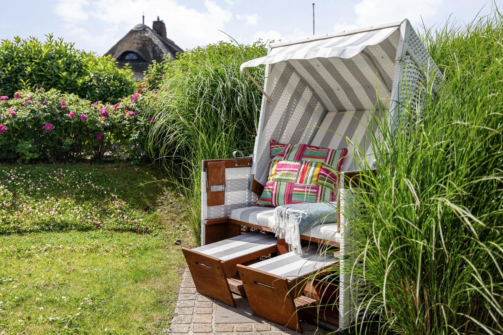 Hochwertiger Strandkorb mit buntem Kissen und Decke im Garten. Hintergrund: Haus mit Schilf und Rosen.