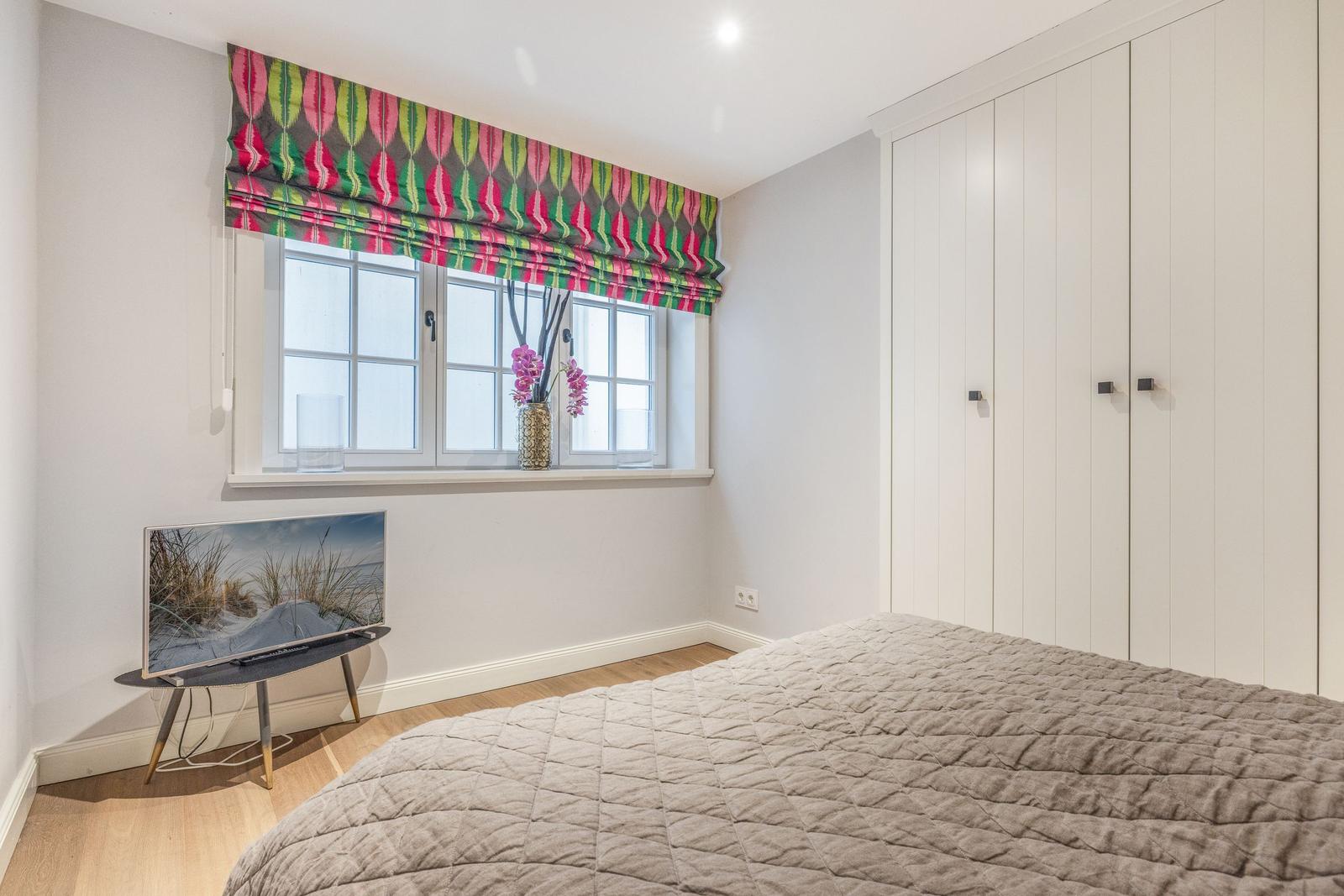 Schlafzimmer mit Bett, Kleiderschrank und Fenster mit buntem Rollladen.