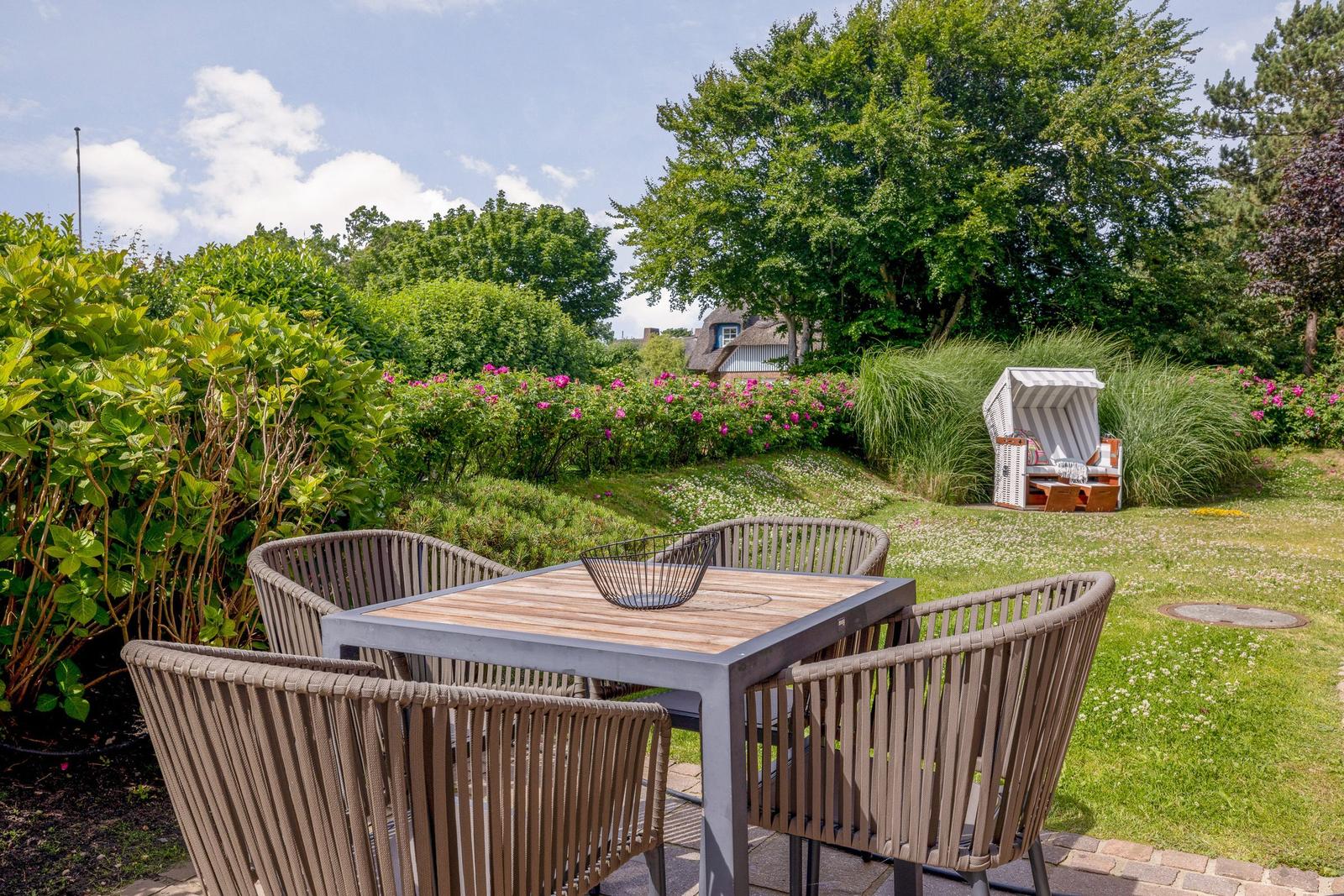 Terrasse mit Tisch und Stühlen, Blick auf Garten mit Blumen und Strandkorb.