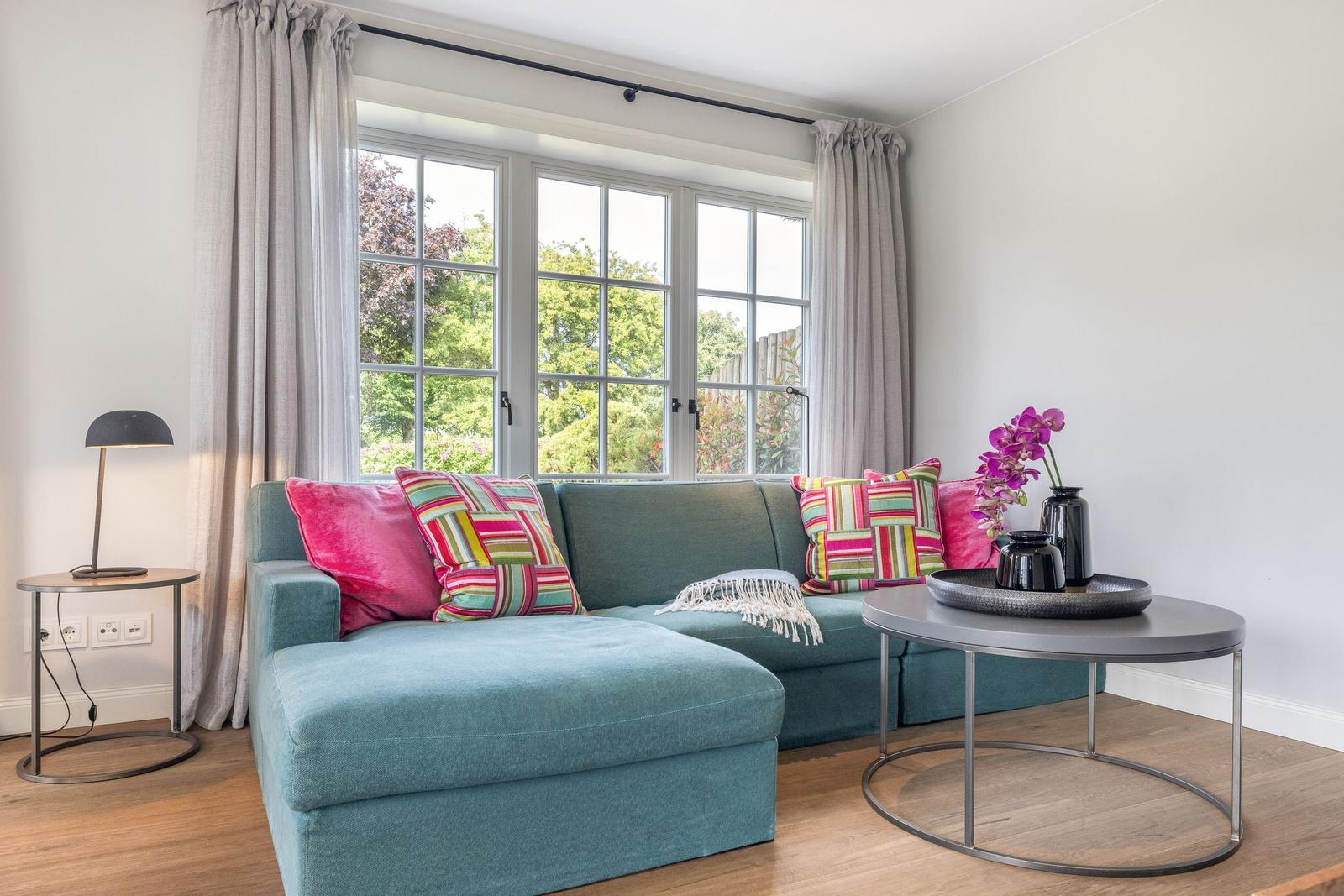Ein Wohnzimmer mit grünem Sofa, bunten Kissen und großem Fenster mit Blick auf den Garten.