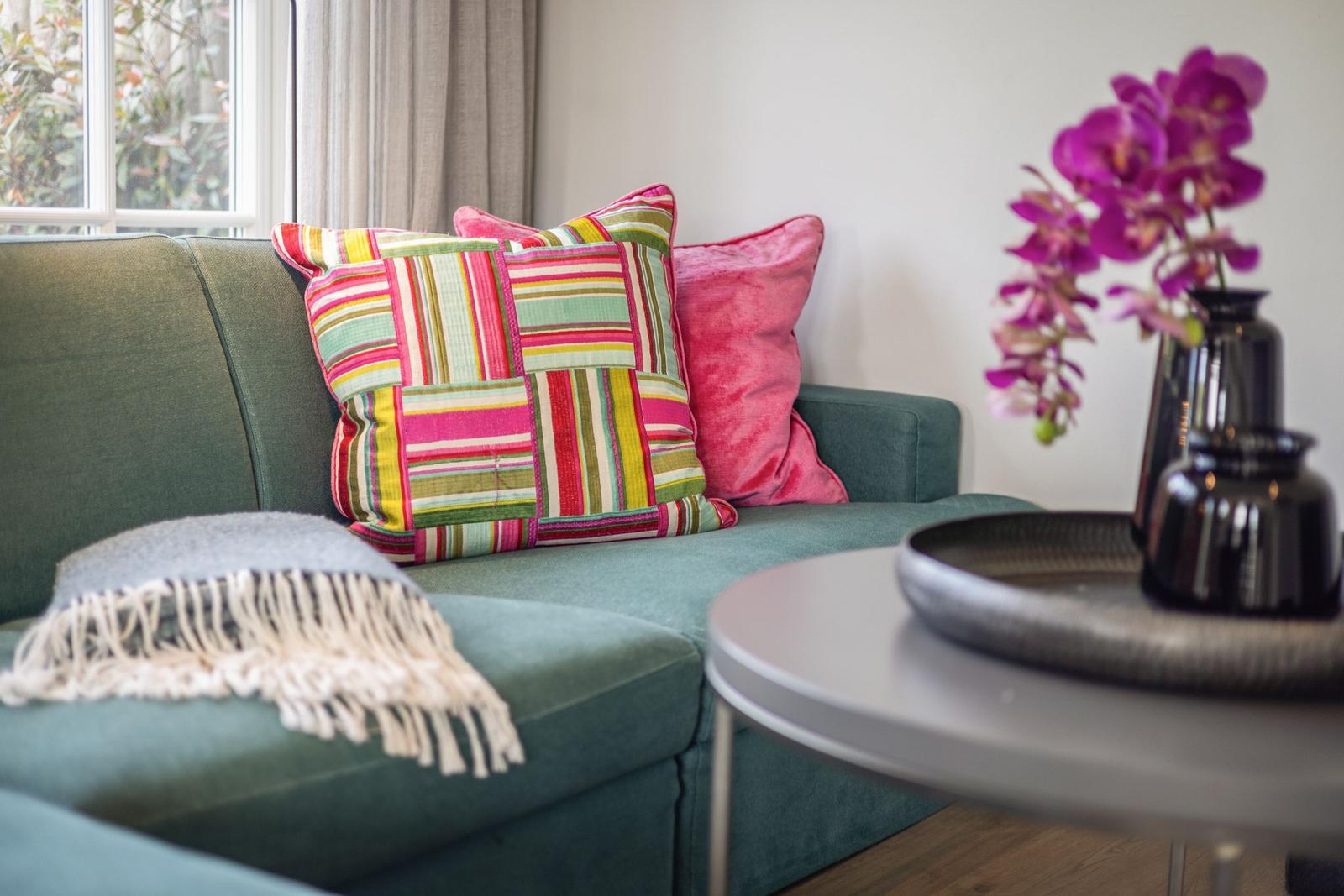 Ein grüner Sofa mit bunten Kissen und einer Decke. Auf einem Tisch steht eine Orchidee.
