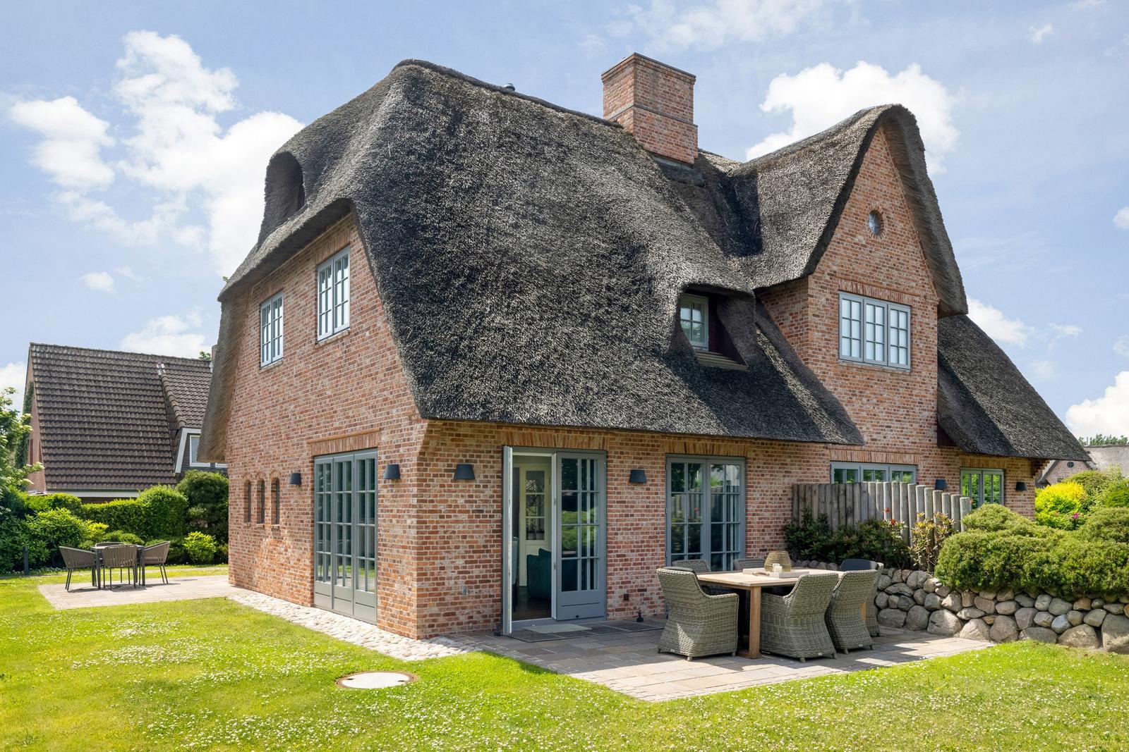 Haus mit Strohdach, Terrasse und Garten. Holztische und Stühle im Freien.