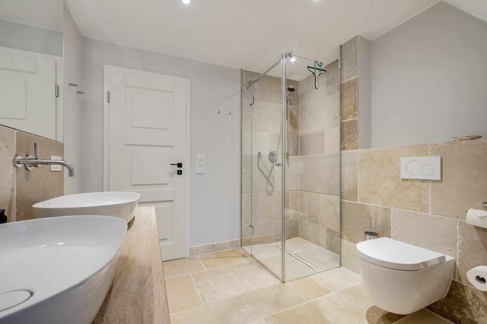 Badezimmer mit Dusche, Toilette und zwei Waschbecken auf Holzplatte.