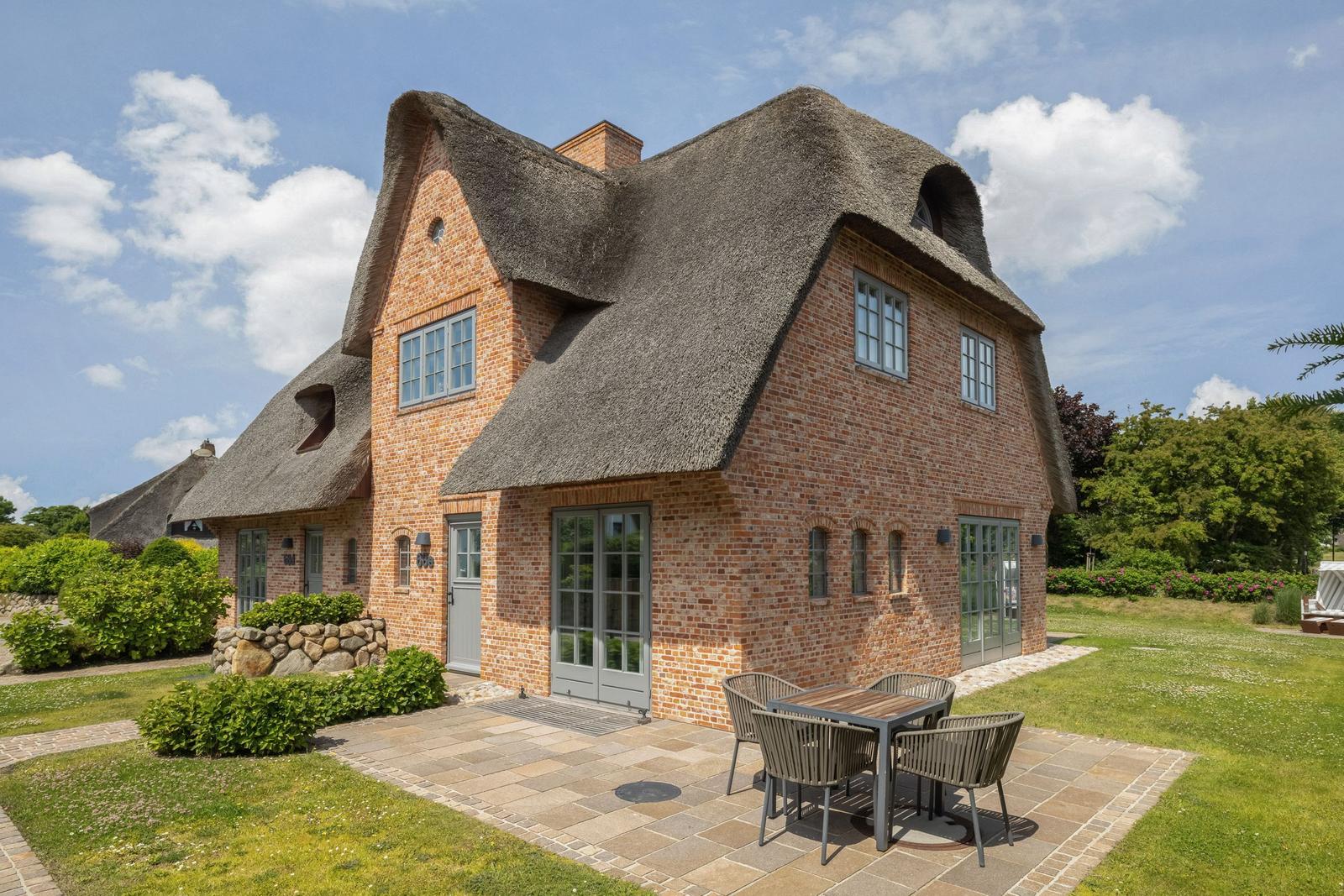 Haus mit Strohdach, Steinplattenhof und Garten. Tisch und Stühle auf der Terrasse.