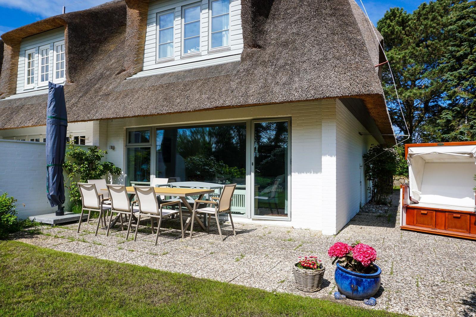 Haus mit Strohdach, Terrasse mit Tisch und Stühlen, Blumenkübel und Gartenbereich.
