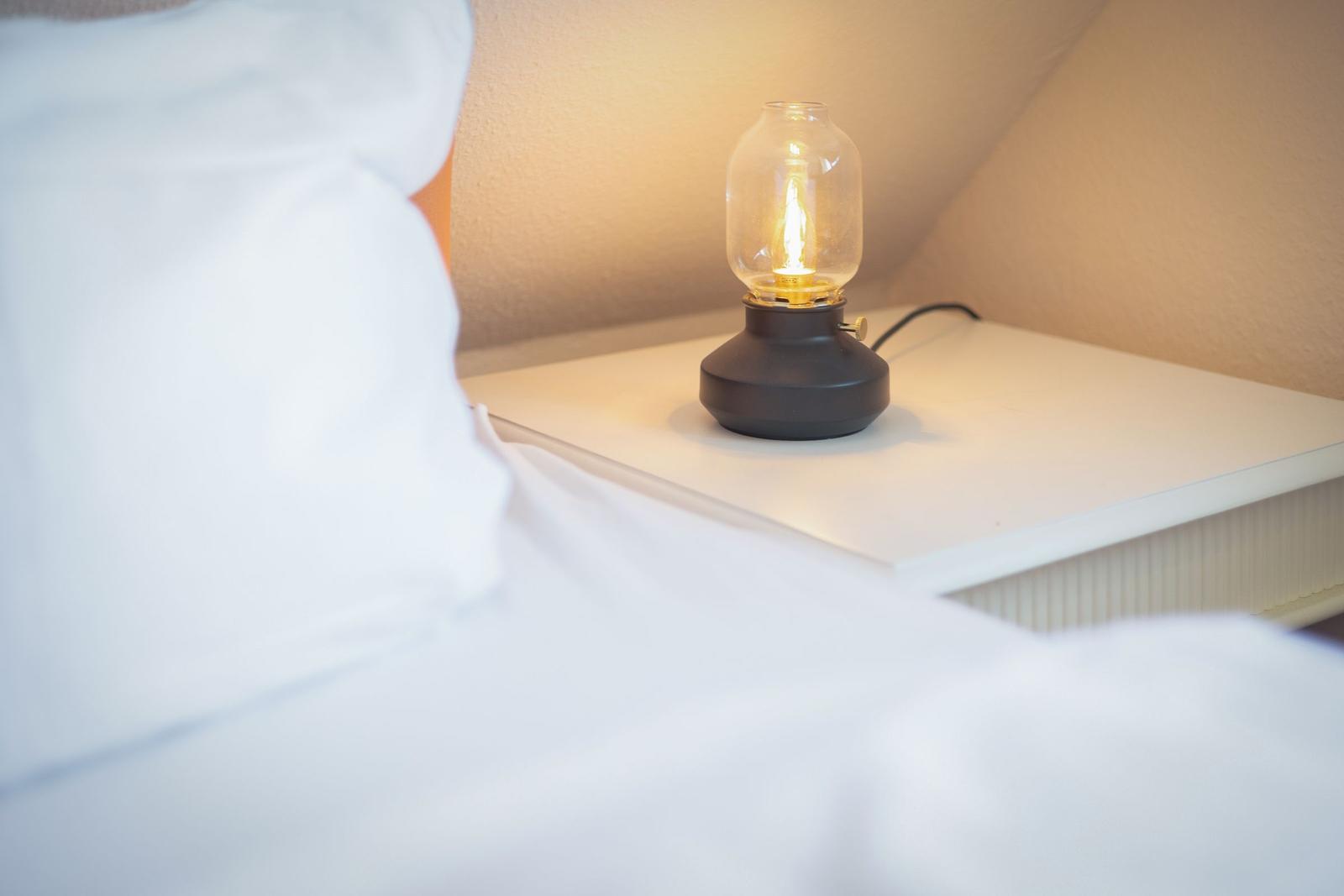 A lamp with a glass housing sits on a nightstand next to a bed with white linens.