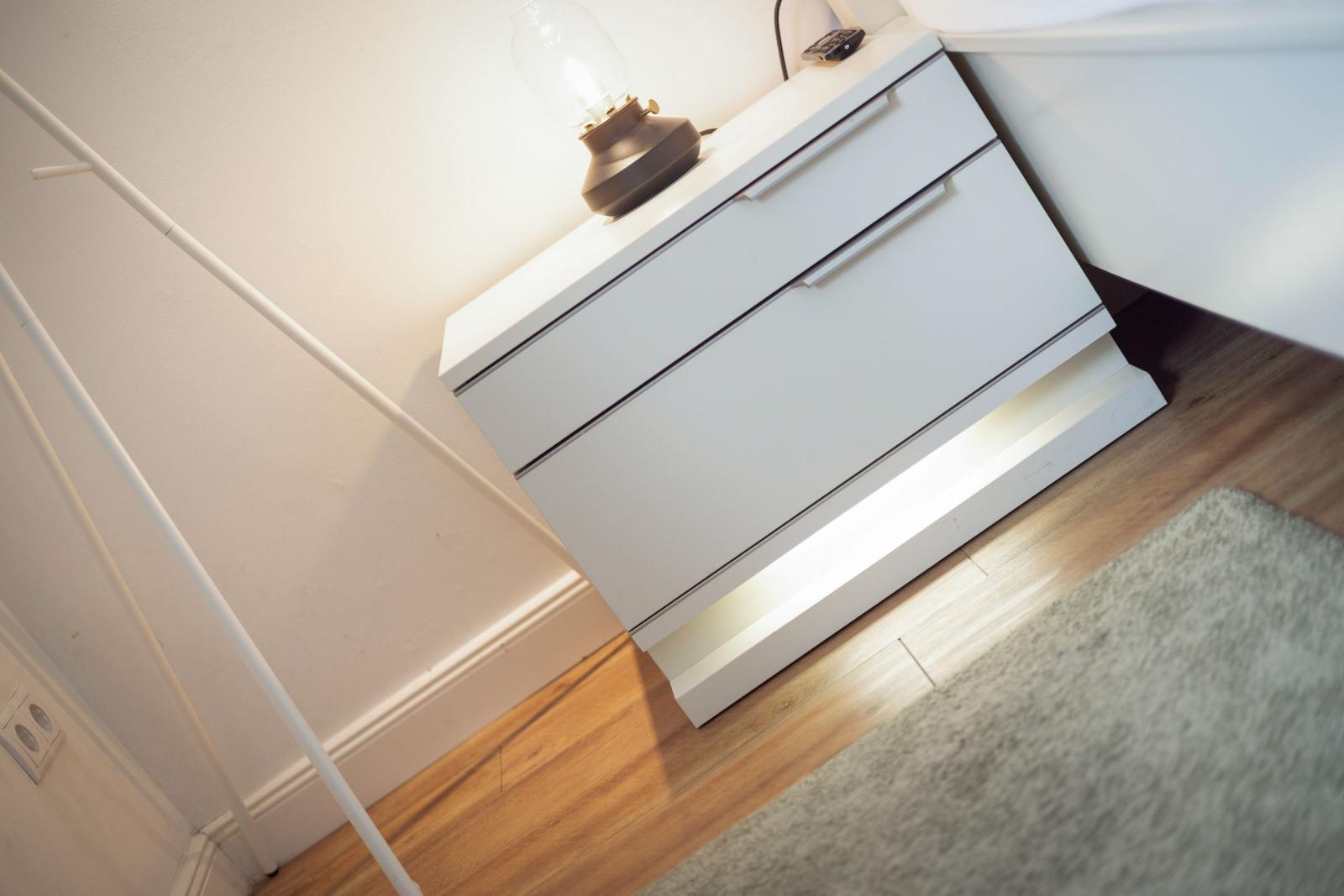 White nightstand with three drawers, lamp, and remote. Wooden floor and rug.