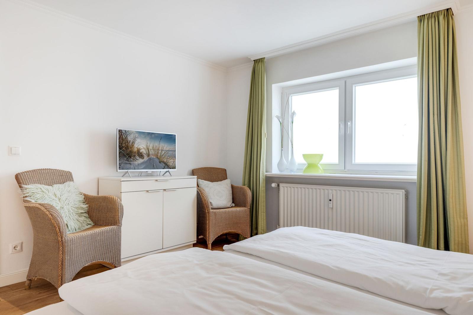 Bedroom with bed, two chairs, TV, and window with green curtains.
