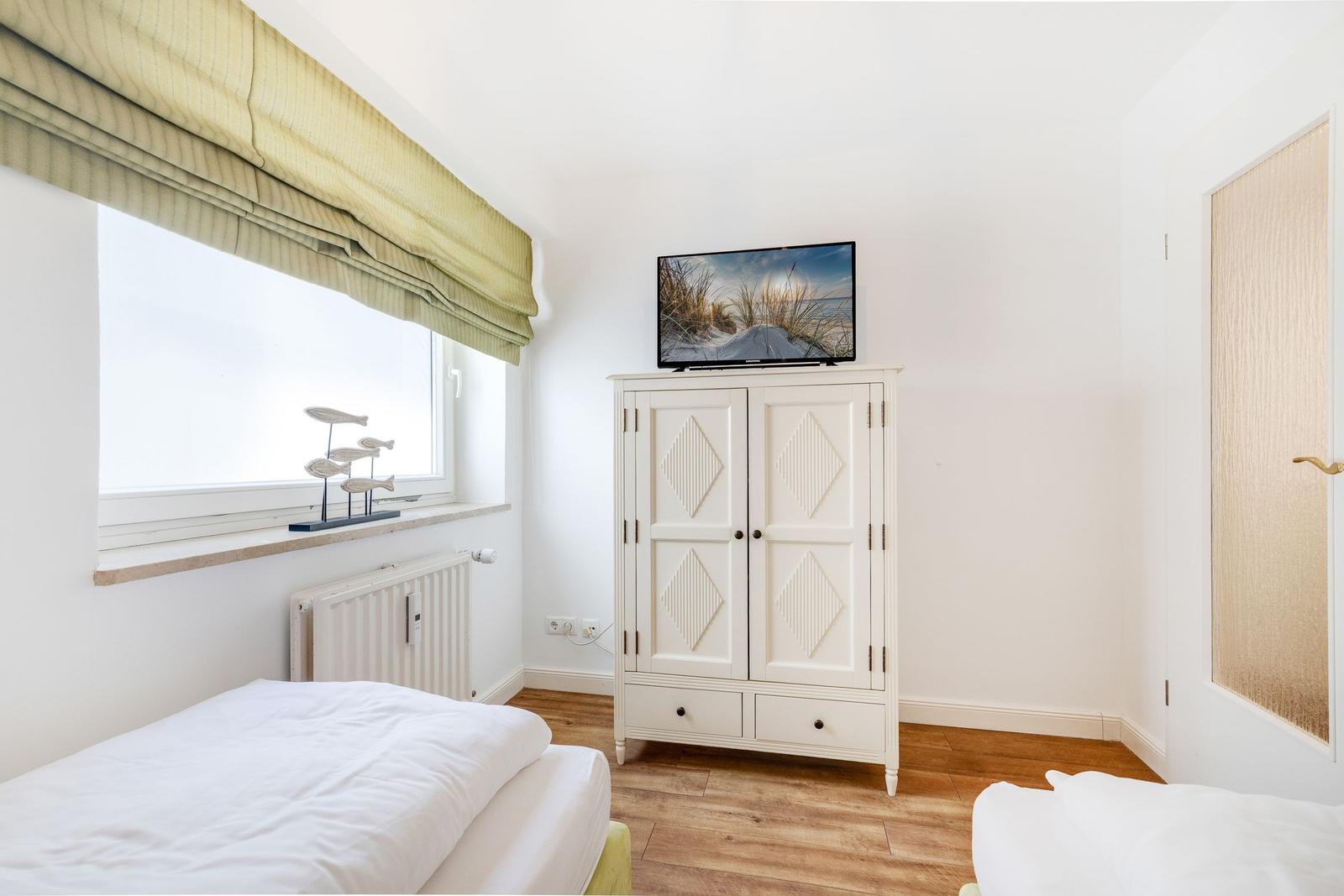 Room with two beds, white wardrobe, and TV. Window with roller blind and floor heating.