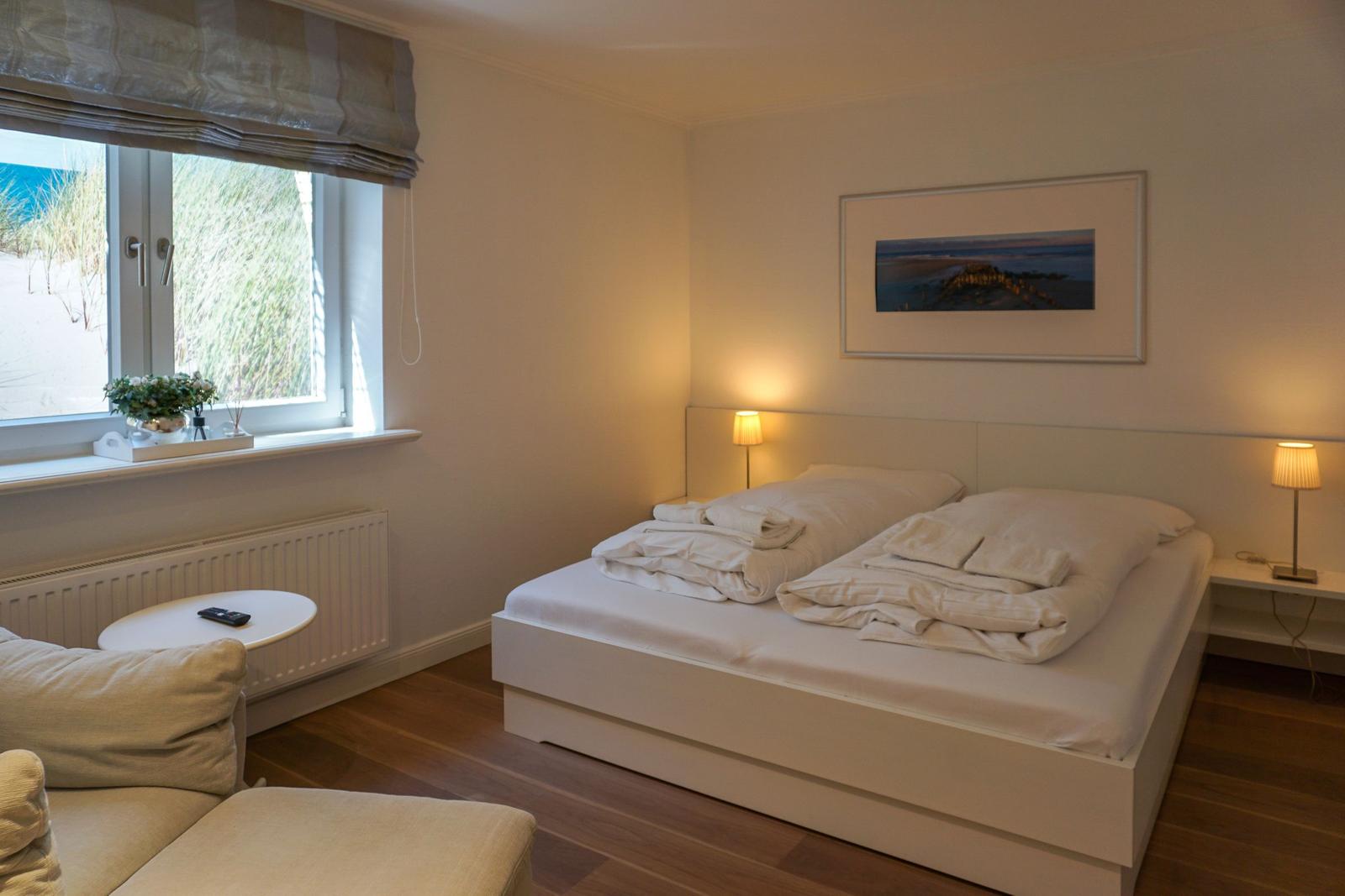 Schlafzimmer mit Bett, Nachttisch und Fensterblick auf Strand.