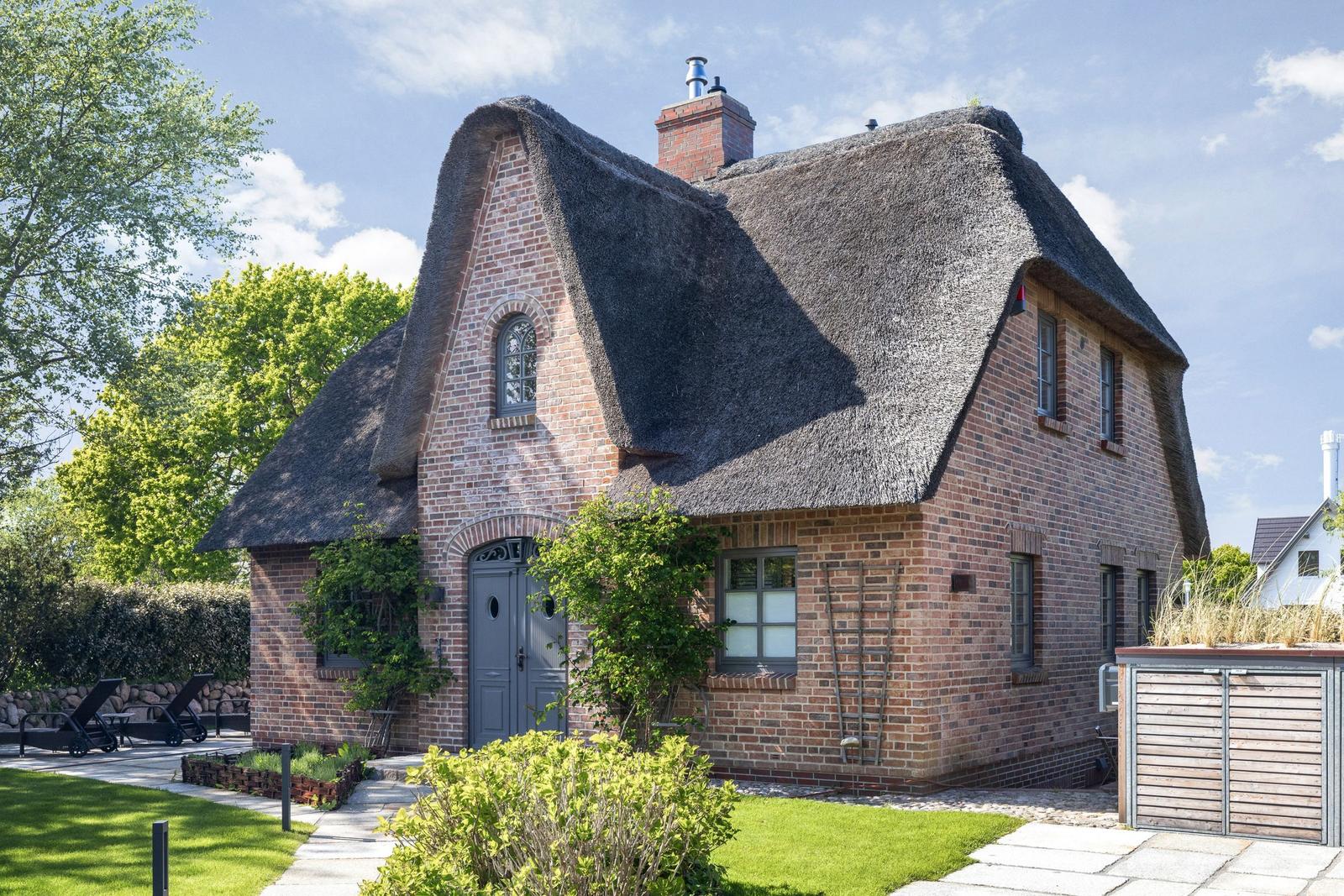 Ein Haus mit strohgedecktem Dach und Backsteinfassade. Gartenbereich mit Liegestühlen.