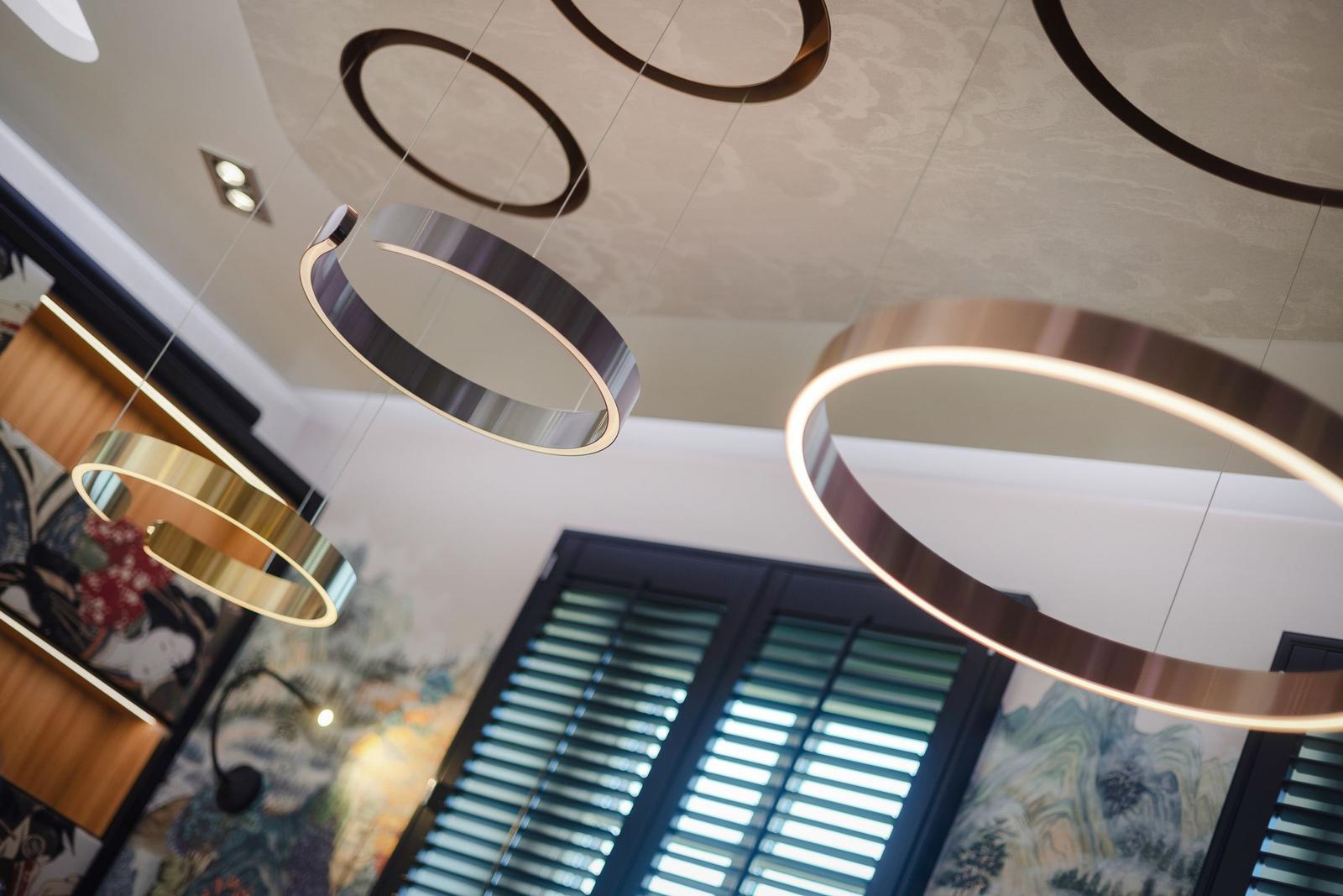 Deckenleuchten mit Ringen hängen über einem Raum mit Jalousien und Wandbildern.