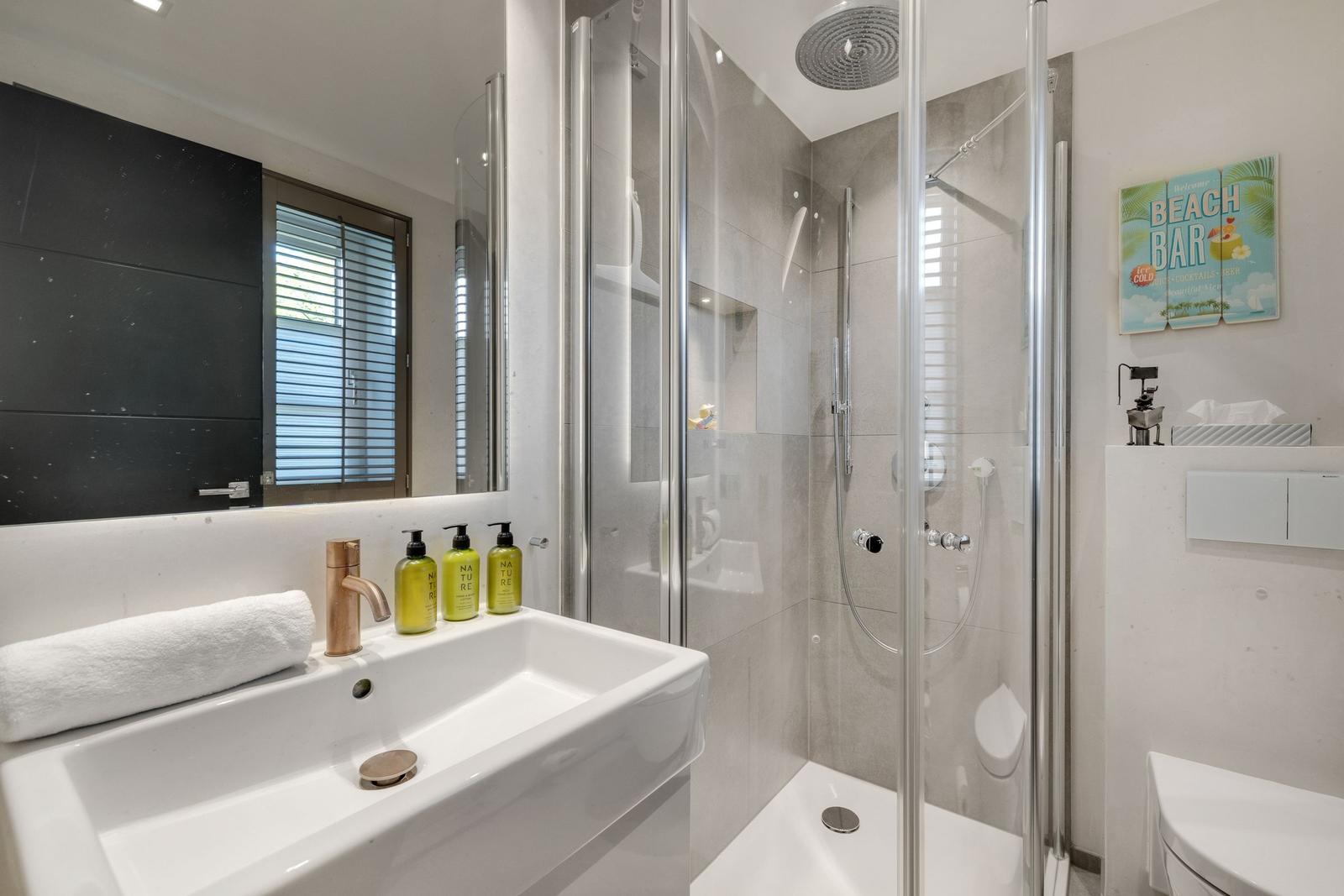 Badezimmer mit Dusche, Waschbecken und Spiegel. Auf Wand hängt ein Strandposter.