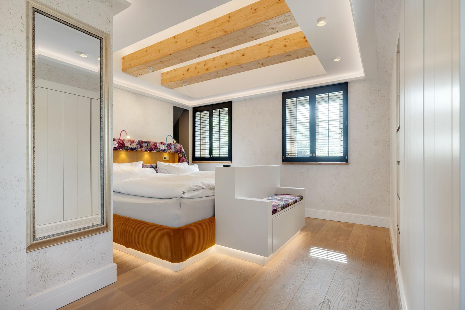 Schlafzimmer mit Bett, Fenstern und Holzboden. Deckenbalken und Spiegel sichtbar.