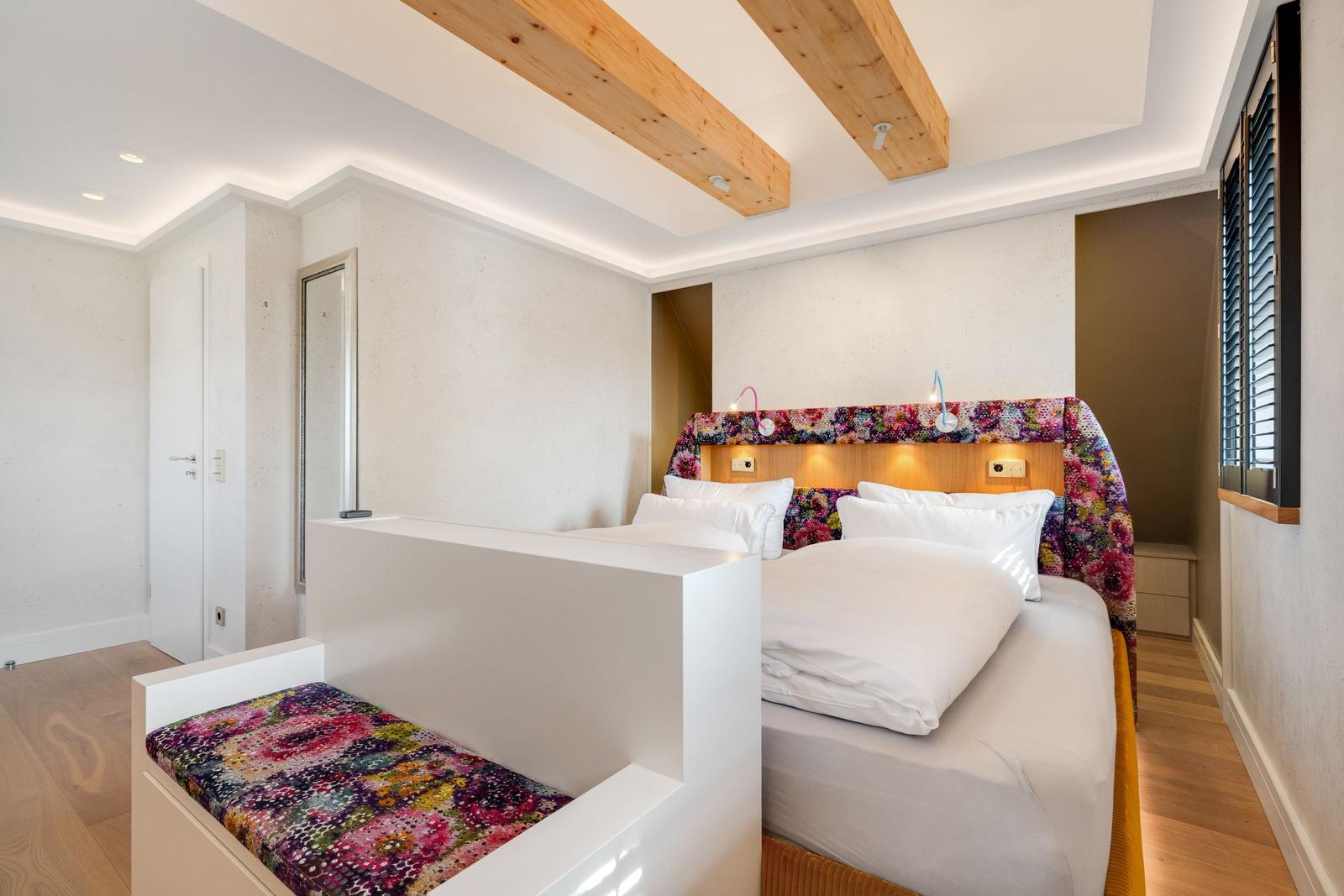 Schlafzimmer mit Bett, Kopfteil und Sitzbank mit floraler Tapete. Holzbalken an der Decke.
