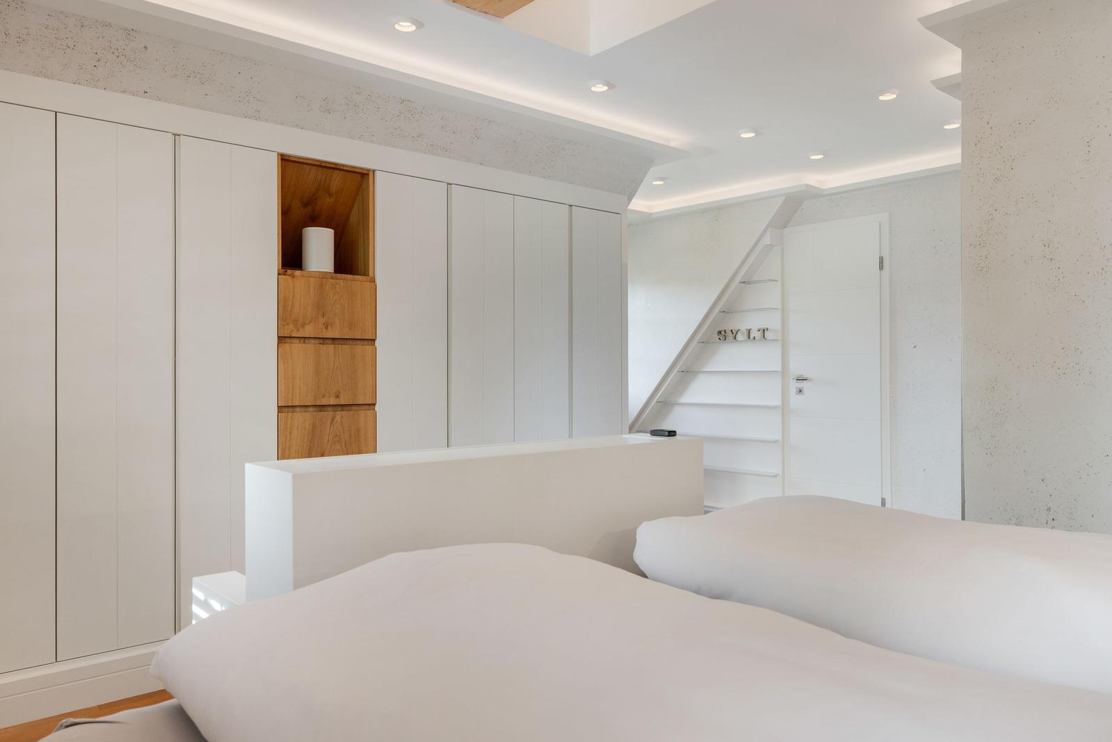 Schlafzimmer mit weißem Bett, Schrank und Treppe zur oberen Etage.