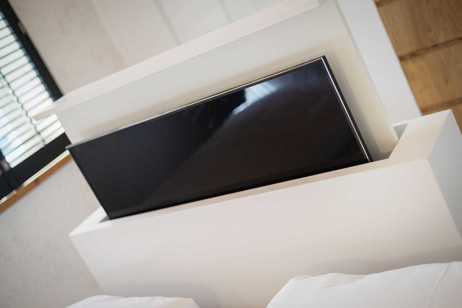 Flachbild-TV in weißem Möbelstück, daneben Fenster mit Jalousie und Bett