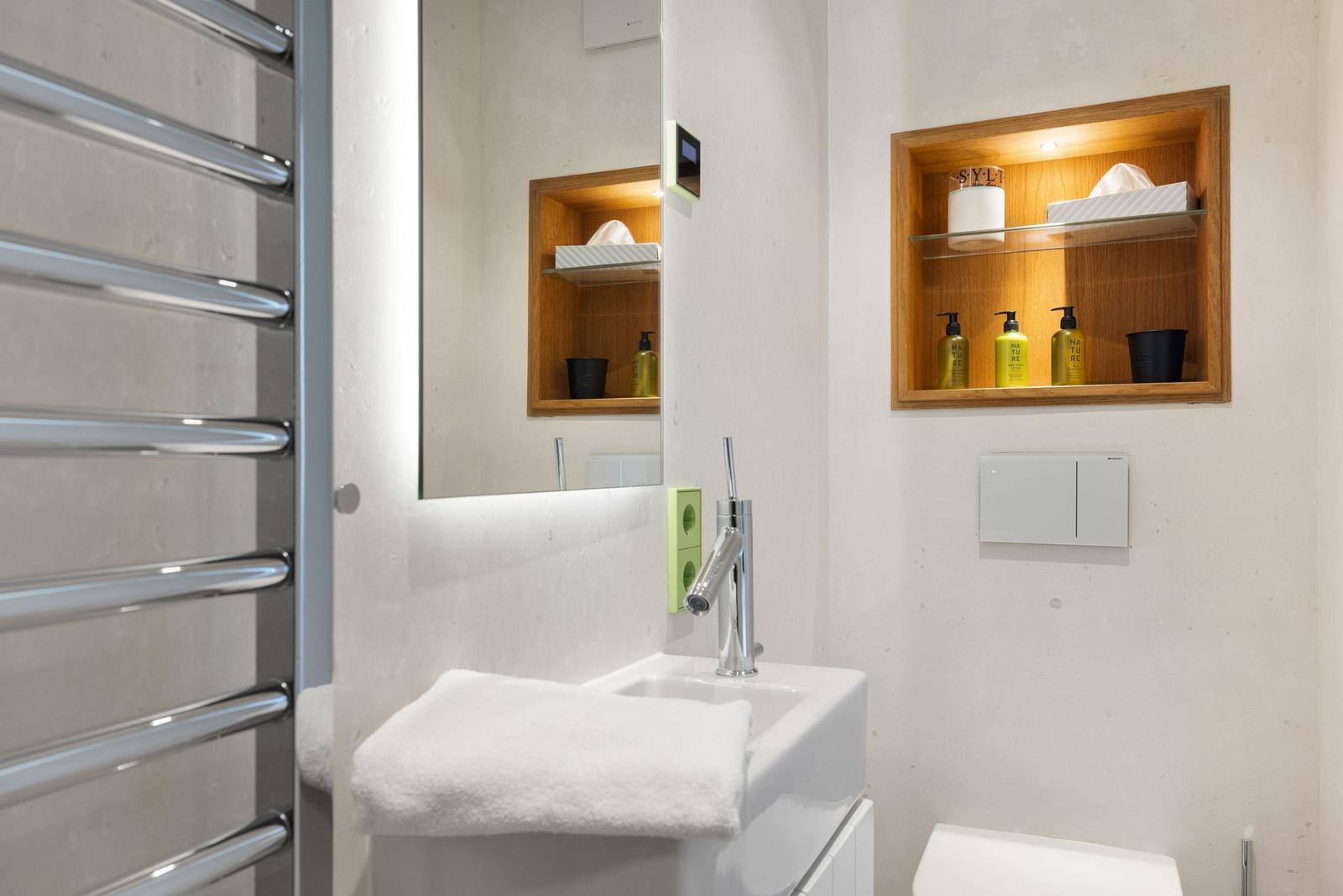 Badezimmer mit Waschbecken, Spiegel und eingebauter Holzregalwand mit Pflegeprodukten.
