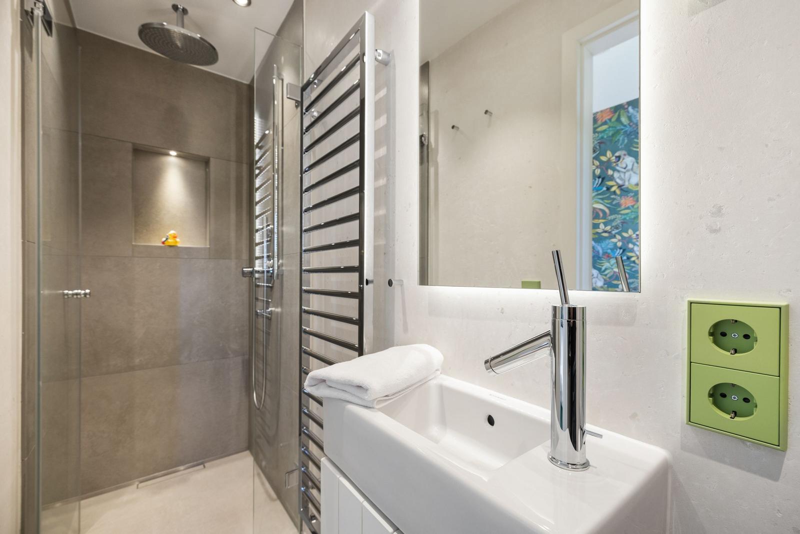 Moderne Dusche mit Regenduschkopf und Handtuchhalter. Waschbecken mit Spiegel und grüne Steckdose.