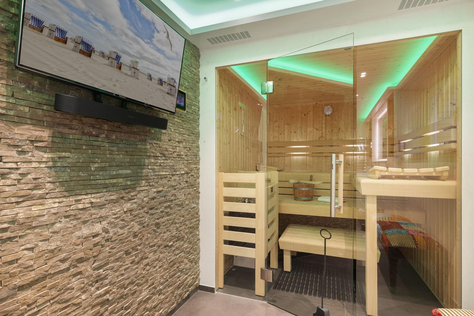 Sauna mit Holzbank und Glaswand. An der Wand hängt ein Fernseher mit Strandbild.