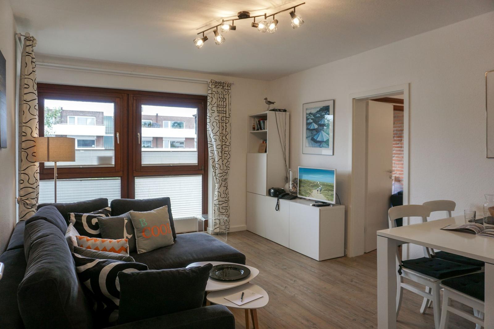 Wohnzimmer mit Sofa, Fernseher und Essbereich. Fenster mit Vorhängen und Holzboden.