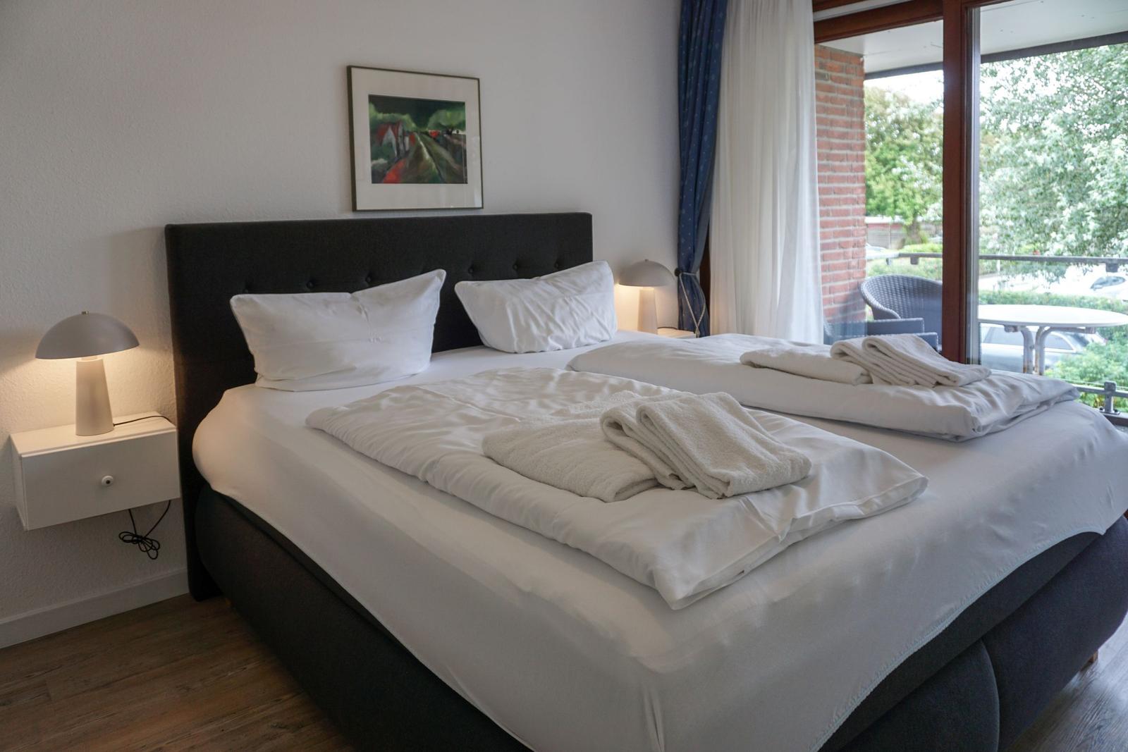 Doppelbett mit weißen Bettwäsche und Handtüchern. Fensterblick auf Balkon und Grünfläche.
