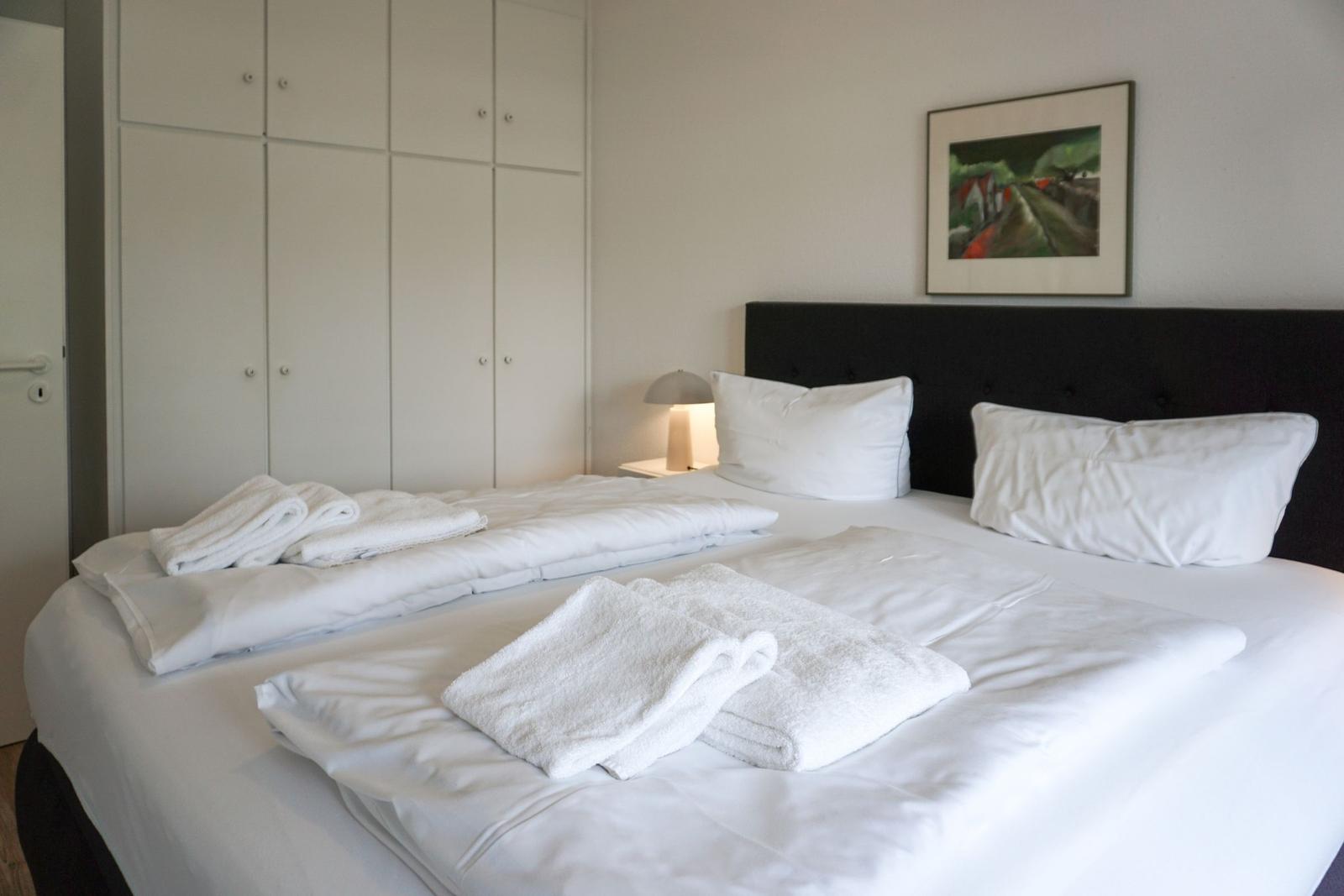 Doppelbett mit weißen Bettwäsche und Handtüchern. Hintergrund: Kleiderregal und Wandbild.