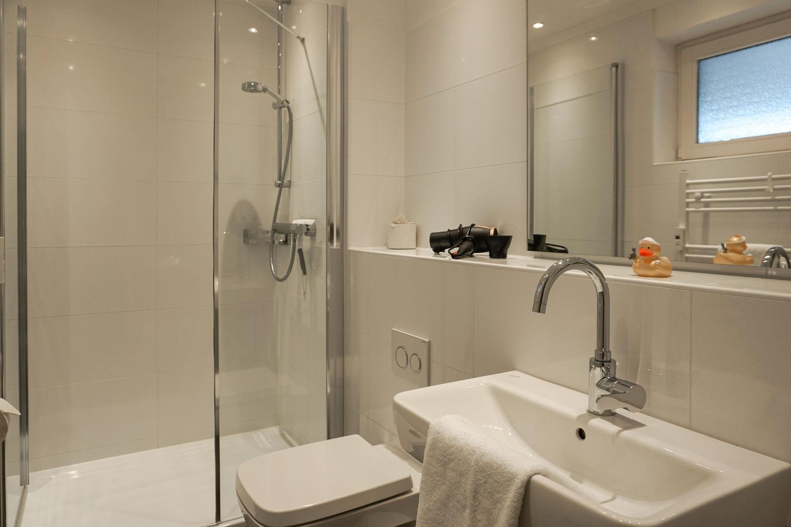 Badezimmer mit Dusche, Waschbecken und Toilette. Ein gelber Enten-Spielzeug ist auf dem Waschbecken.