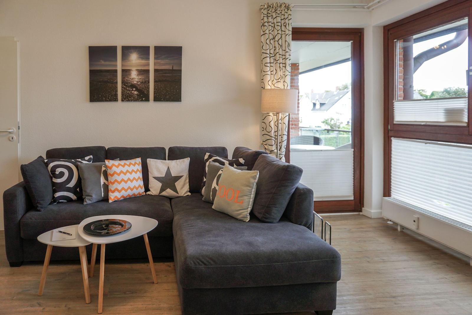 Ein Wohnzimmer mit grauem Sofa, Kissen und einem runden Tisch. An der Wand hängt ein Bild.