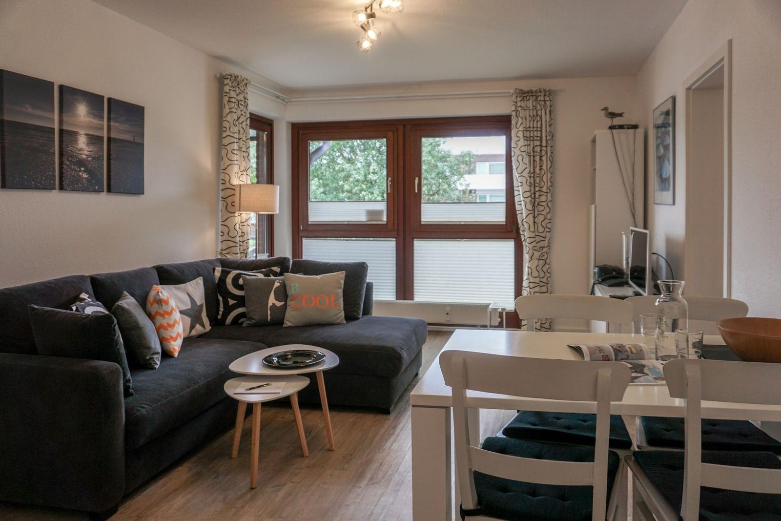 Wohn- und Essbereich mit Sofa, Tisch und Fenster. Möbel sind weiß und dunkel.