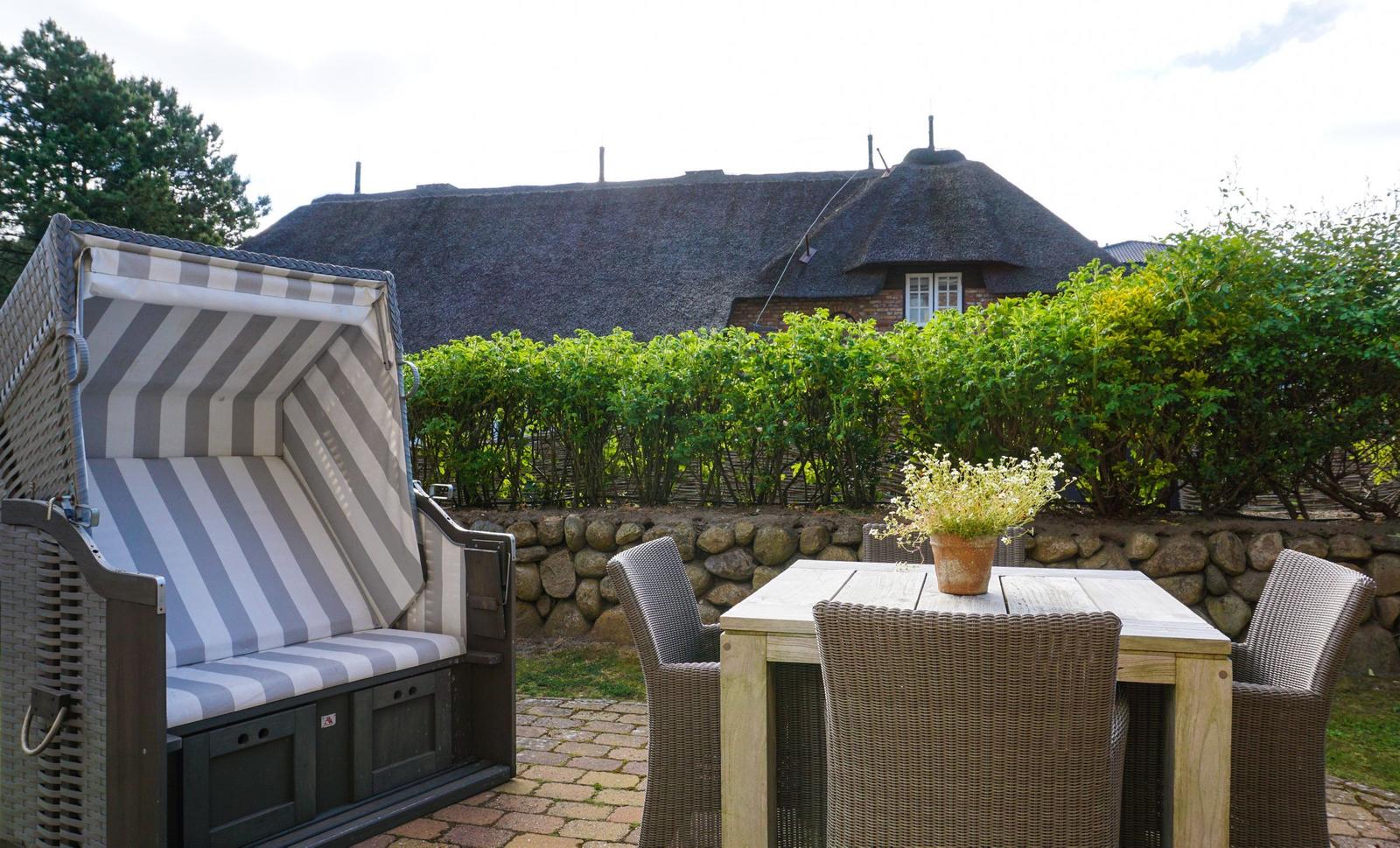 Terrace with beach chair, table and chairs. Backyard with stone wall and green hedge. Roof made of straw.
