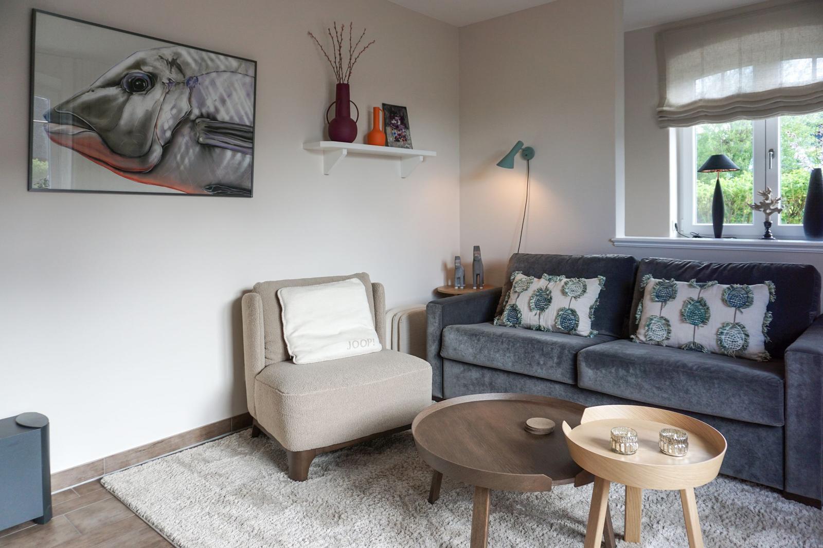 A living room with a gray sofa, beige armchair, and round tables. A large fish painting hangs on the wall.