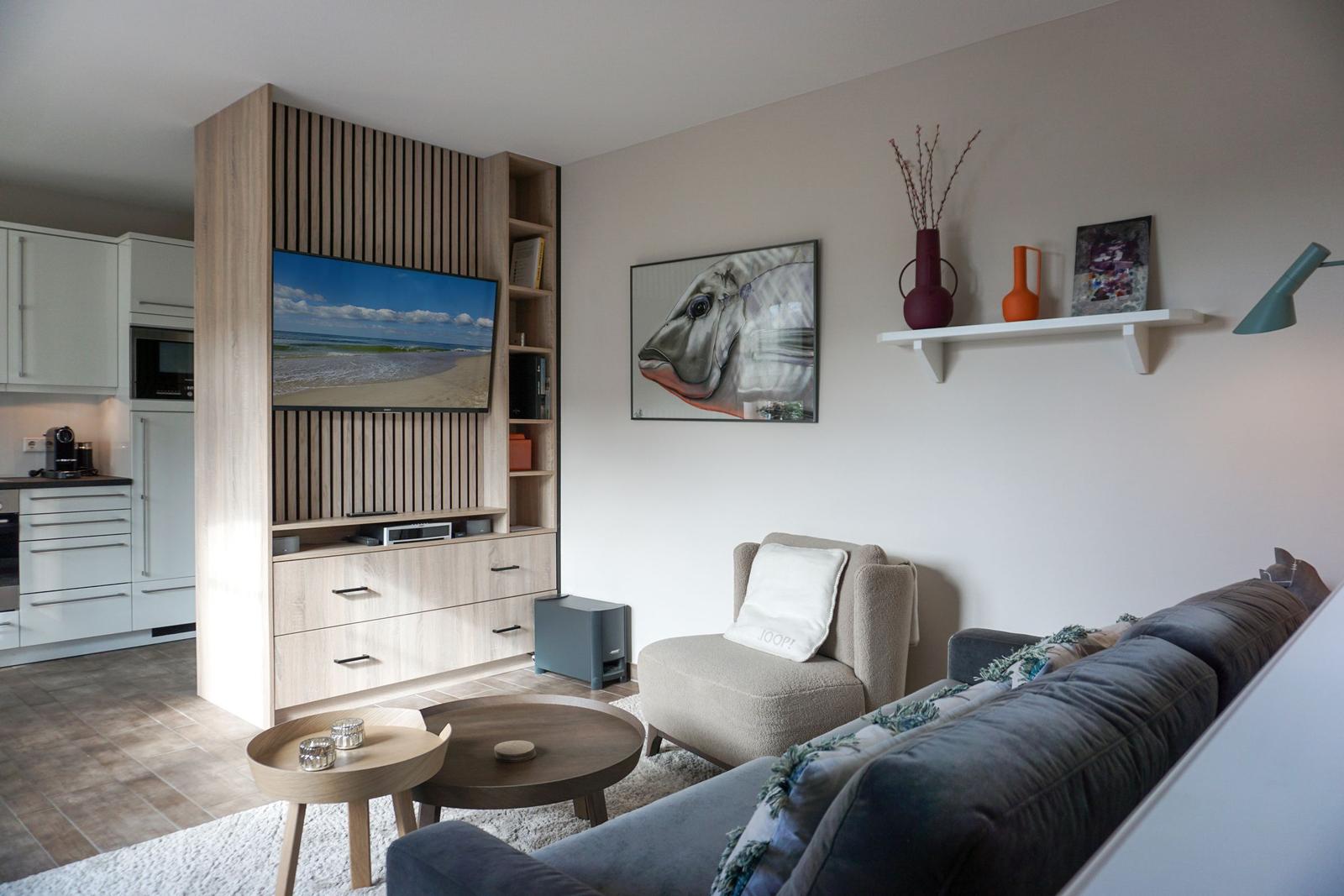 Living room with sofa, armchair, and TV in wooden unit. Kitchen in background.