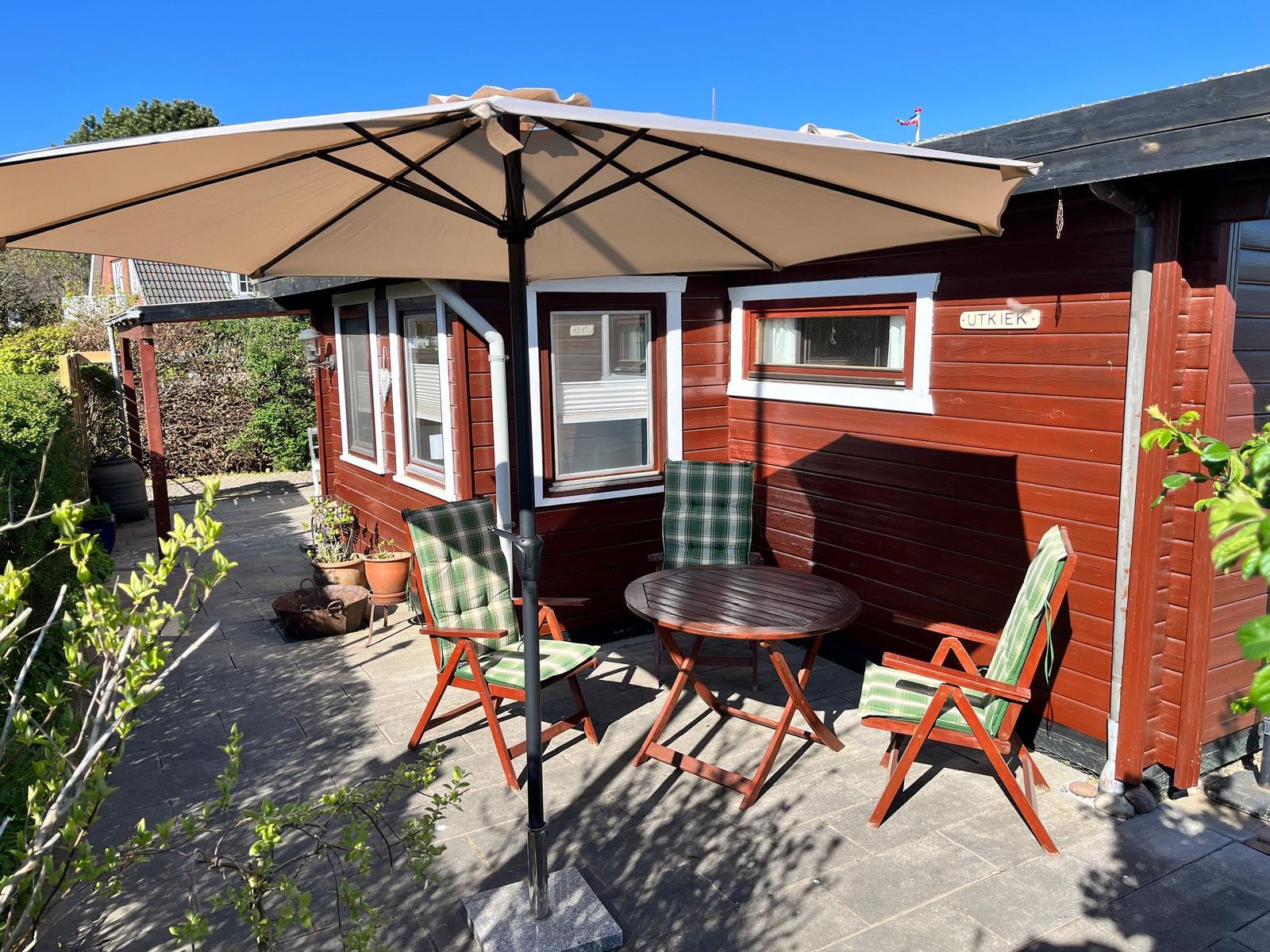 Terrasse mit Tisch, drei Stühlen und Sonnenschutz vor rotem Holzhaus.