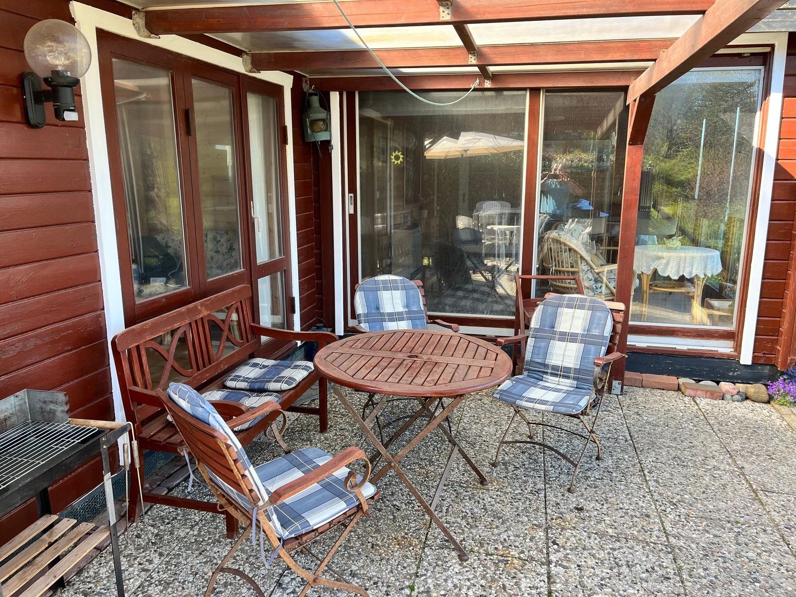 Terrasse mit Holztisch, Stühlen und Grill. Glasfront führt ins Haus.