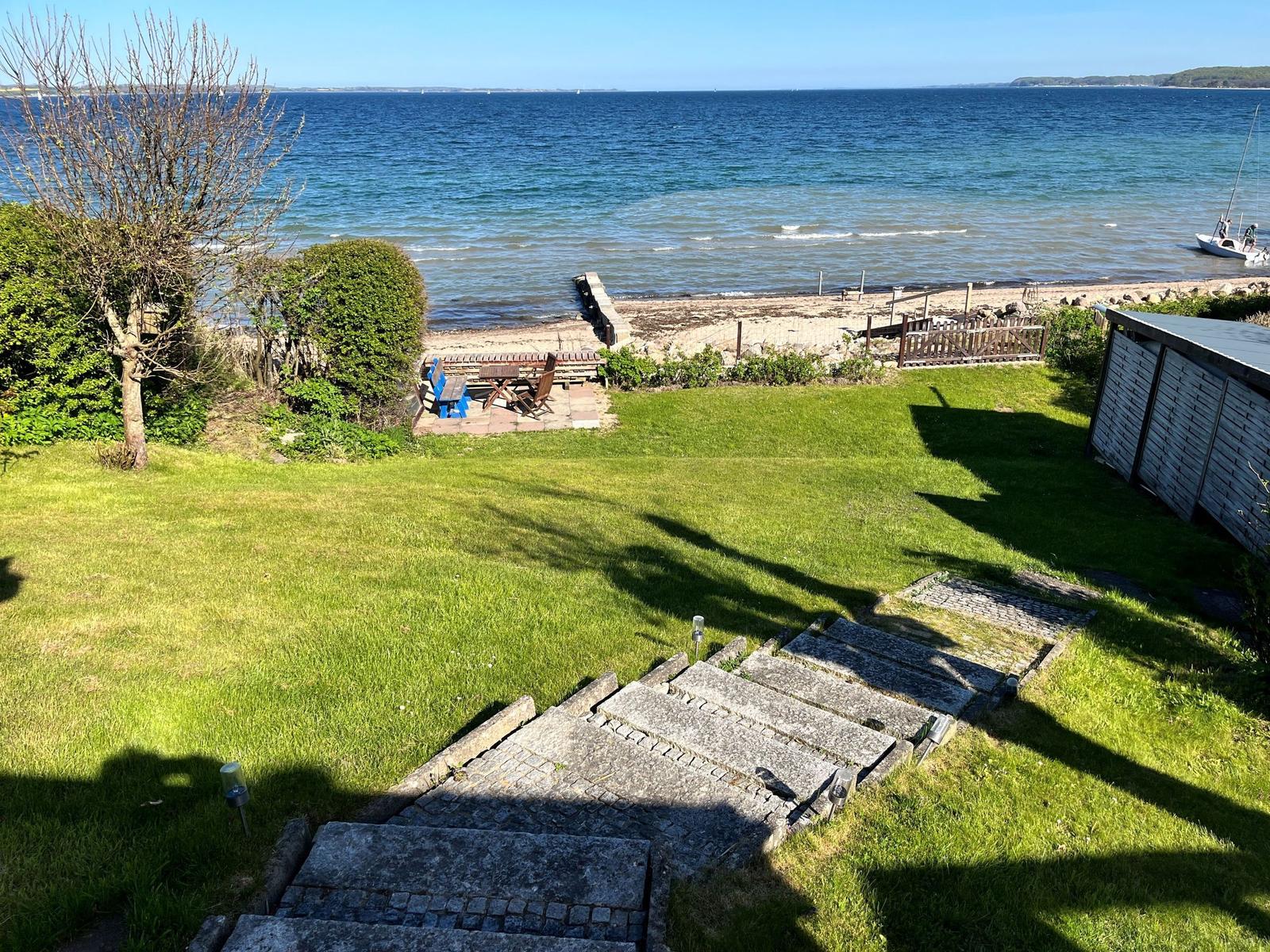 Grüner Garten mit Steinstufen führt zum Strand. Holztisch, Stühle und Bootssteg sind sichtbar.