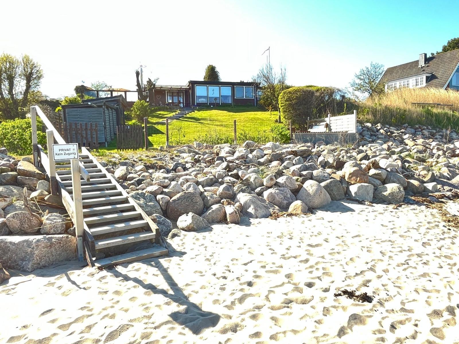 Strand mit Steinschutz, Holzstufen und modernem Haus auf grünem Hang.