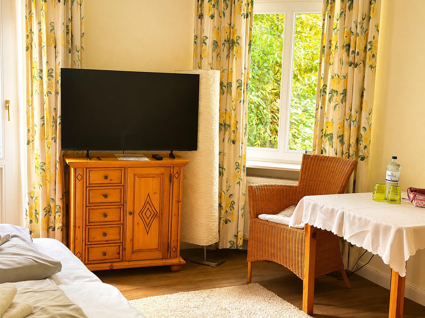 Ein Zimmer mit Bett, TV-Schrank, Stuhl und Tisch. Fenster mit gelben Vorhängen.